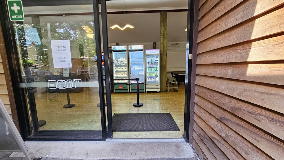 Entryway leading to the inside of the Experiences Hut. On top of the left automatic sliding door, a first aid logo and a sign that indicates that the Lone Pine gift shop is located inside are noticeable. Upon entering, a black floormat and bollards are apparent. The floor surface has timber flooring throughout. Display fridges with snacks are noticeable in the center. The bird-like design of a fluorescent light is prominently hanging on the ceiling.