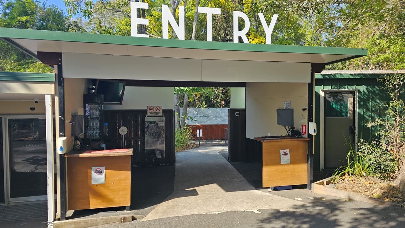 Entryway leading to the ticketing office. A bold “ENTRY” sign is prominent on top of the roof of the cream-structured ticketing office. Two brown desks are apparent with hand sanitizers. Signages are apparent in the entryway. On the ceiling on the left, a TV is apparent, and on the right side is a functional CCTV. Next to the entryway, a green color bond shed is noticeable on the right.