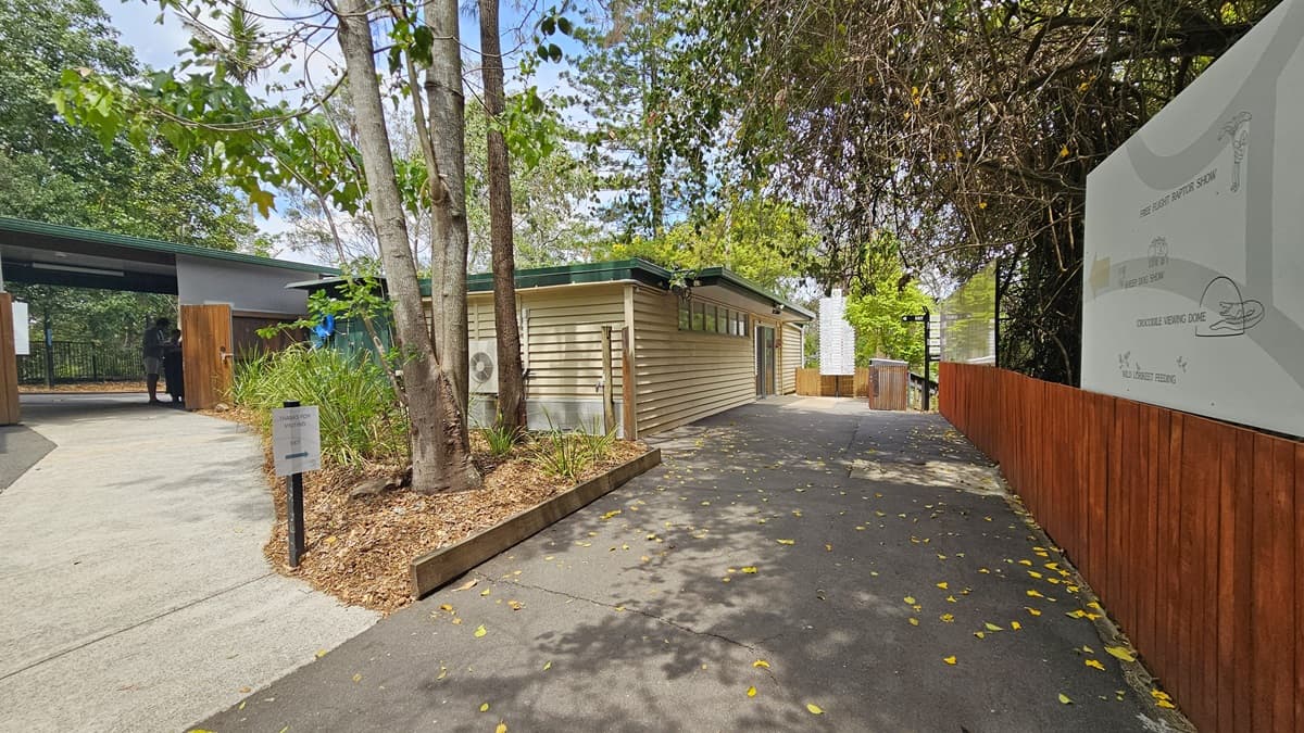 Another pathway inside the sanctuary. Two individuals are visible in the ticketing office situated on the left side. A small signage is placed in the garden filled with plants and trees, between two separate pathways. Behind the garden, the extension of the ticketing office is noticeable. On the right, a lengthy wooden fence and a large sign that indicates various directions of the attractions inside the sanctuary.