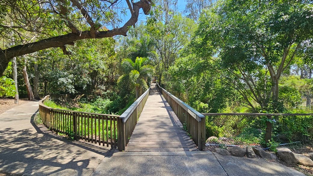 A lengthy foot bridge. The slender structure is adorned with wooden railings and a wooden floor surface. To the left, the wooden barriers clearly fenced a lush section adorned with decorations. On the right, expansive steel railings prominently enclose an area abundant with trees and grass.