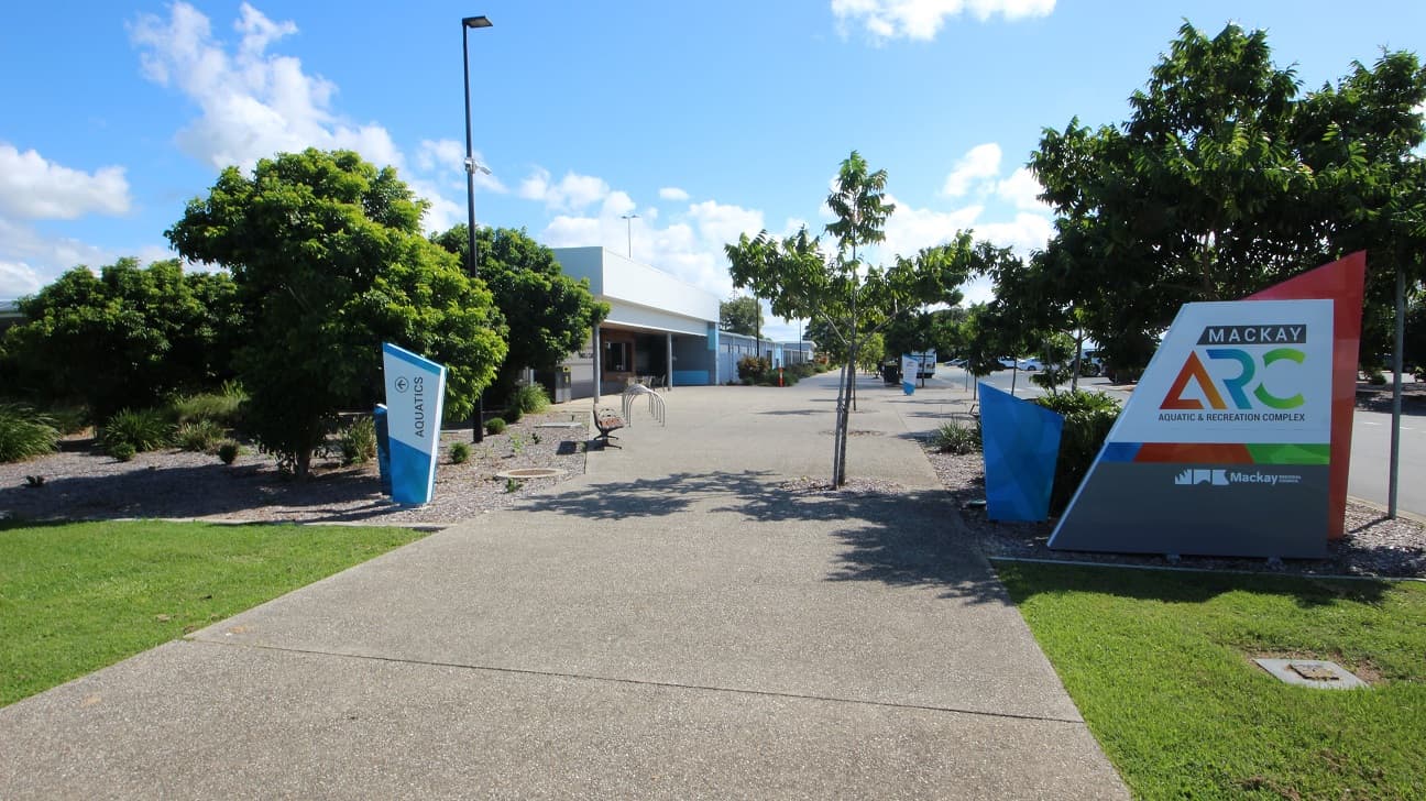 A straight concrete road flanked by green grass and trees. On the right, a concrete structure displays facility signage. Additional concrete signages, including one directing to Aquatics, are adjacent. Trees, street lights, and benches line the left side. Further ahead, a low-set building comes into view.