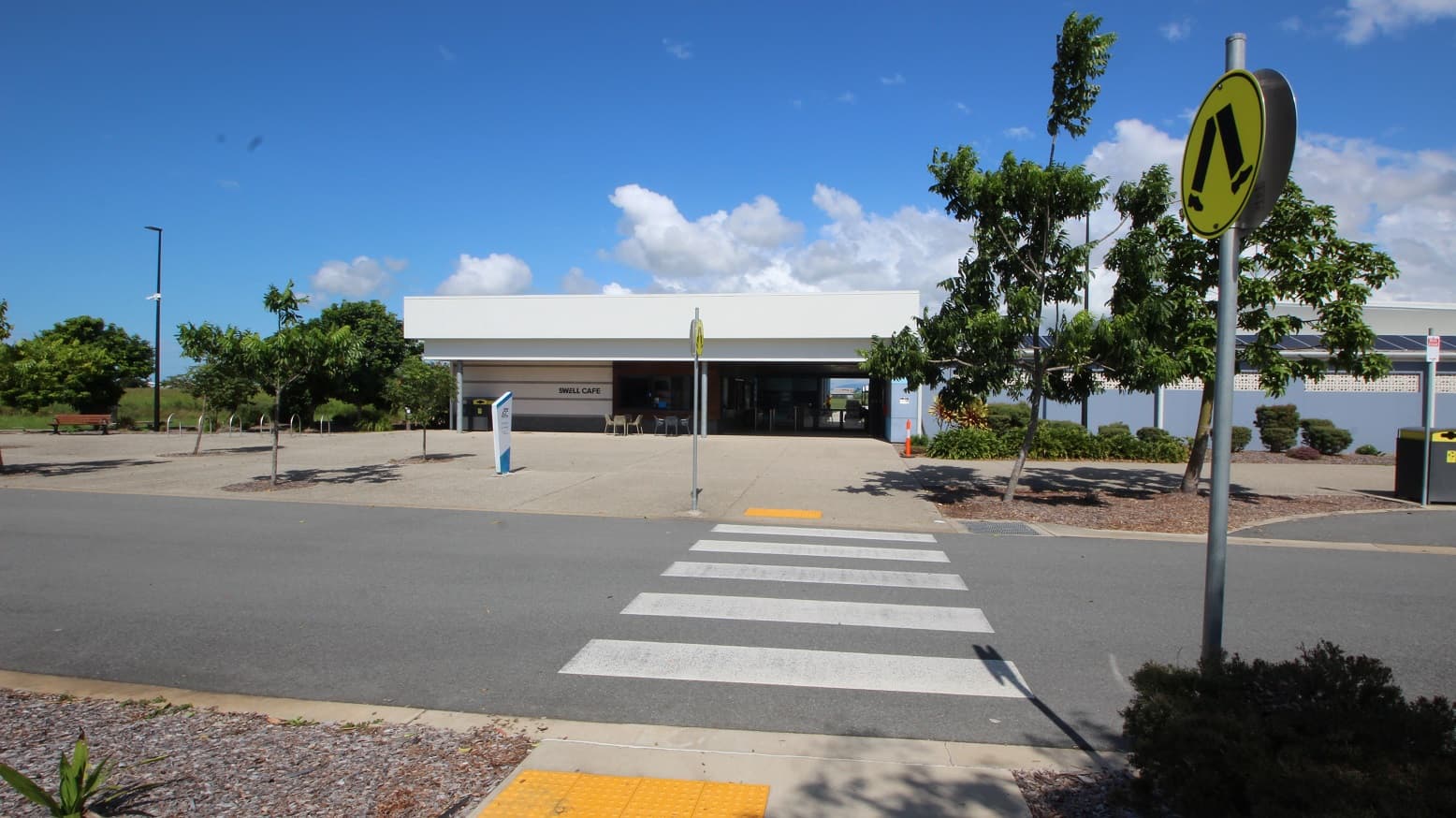 A concrete road with pedestrian markings leads to a low-rise building with a white roof, featuring a step-free ramp. Yellow truncated domes mark both ends of the lane. Road signs line the road edge. Bike racks, a bench chair, and tall street lights are visible on the far left. Trees and trimmed shrubs are on the right.