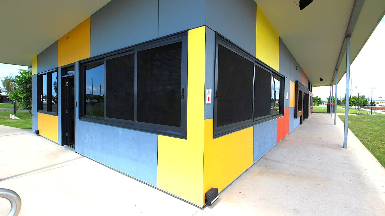 A building with a corner view, featuring a facade of alternating yellow and blue panels. Large, dark-framed windows are set into the walls. The building is situated on a concrete path that extends along its side. The roof is flat and extends outward, supported by metal poles. In the background, there is a grassy area with trees and a fence, and some structures are visible in the distance.