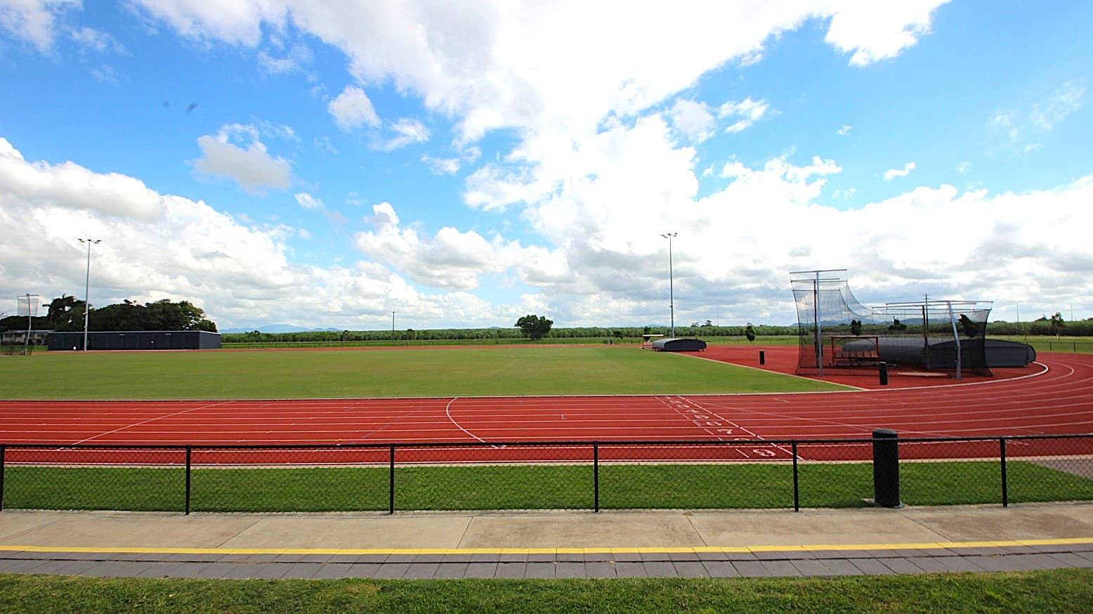 A sports field with a red running track encircling a green grass area. On the right, there is a netted area likely for field events. The track has marked lanes and numbers. In the foreground, a paved path runs parallel to a black metal fence. The background features trees and open fields under a partly cloudy sky.