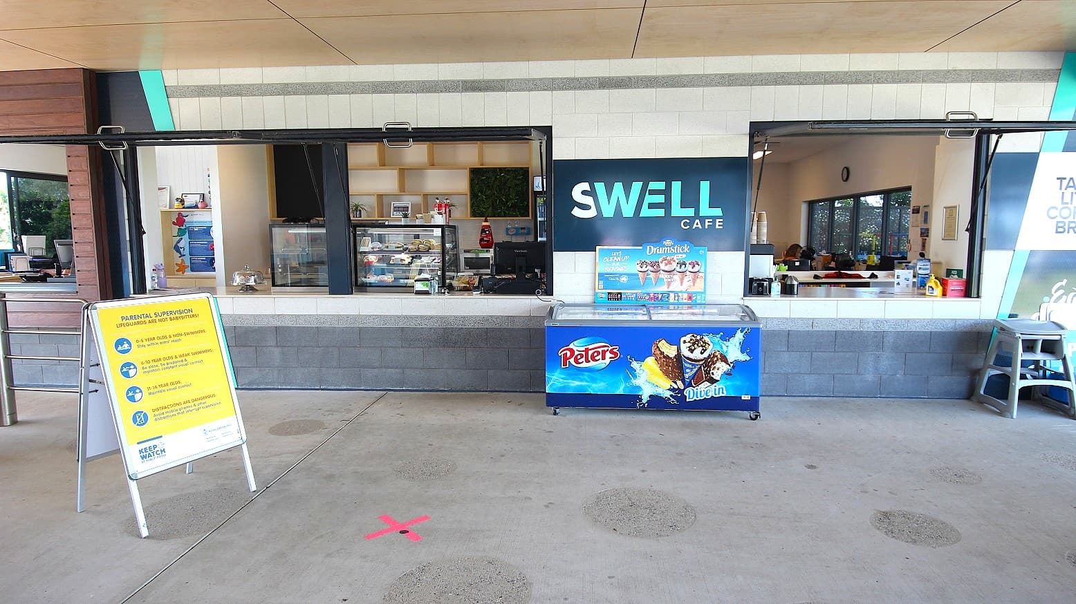 The Swell Cafe displays a grey and white brick facade with a central banner and an ice cream freezer. Inside, food display racks adorn the counter. Wall decorations enhance the ambience. A hot drink dispenser with stacked paper cups is situated on the right. A large banner and a child's high chair are against the wall. A yellow signage is also placed in front.ambi