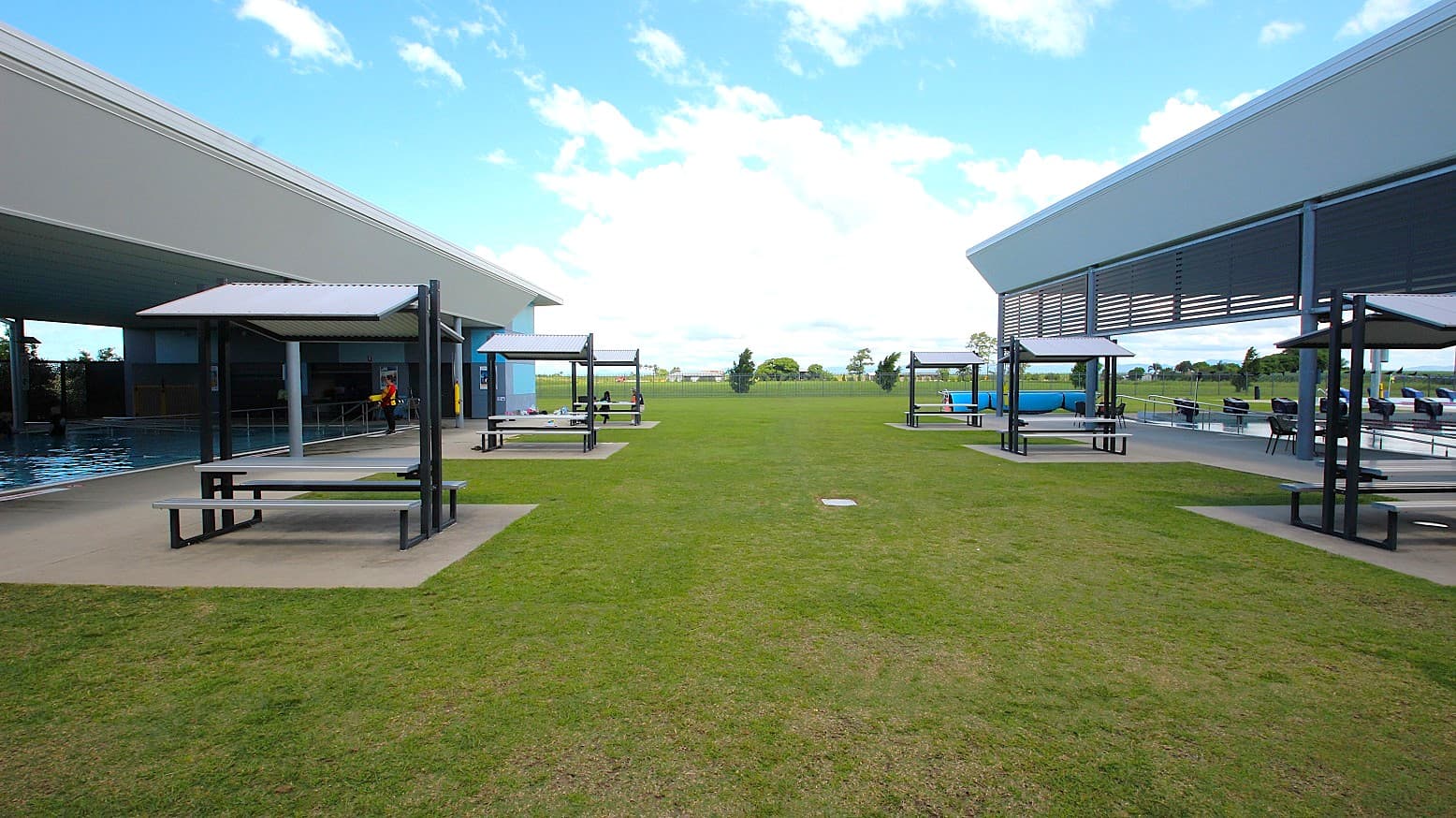 On the left, there is a covered area with a swimming pool and a shaded seating area with benches. On the right, another covered area features similar shaded seating. The middle ground is a grassy open space leading to a distant view of trees and a clear sky. The foreground shows a well-maintained lawn, and the structures have modern, angular designs.