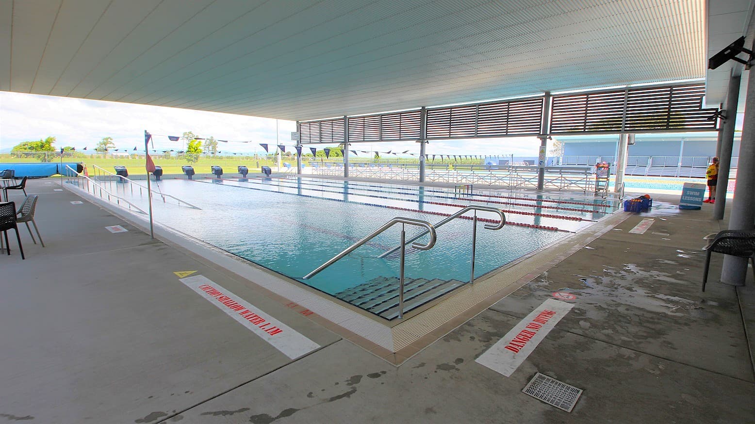 An indoor swimming pool with multiple lanes, bordered by a concrete deck. On the left, there are tables and chairs. The pool has metal handrails leading into the water. The ceiling is covered, and the area is well-lit. In the background, there are large windows with horizontal slats, allowing natural light to enter. Outside, a grassy area and some trees are visible. The floor is wet with visible caution signs.