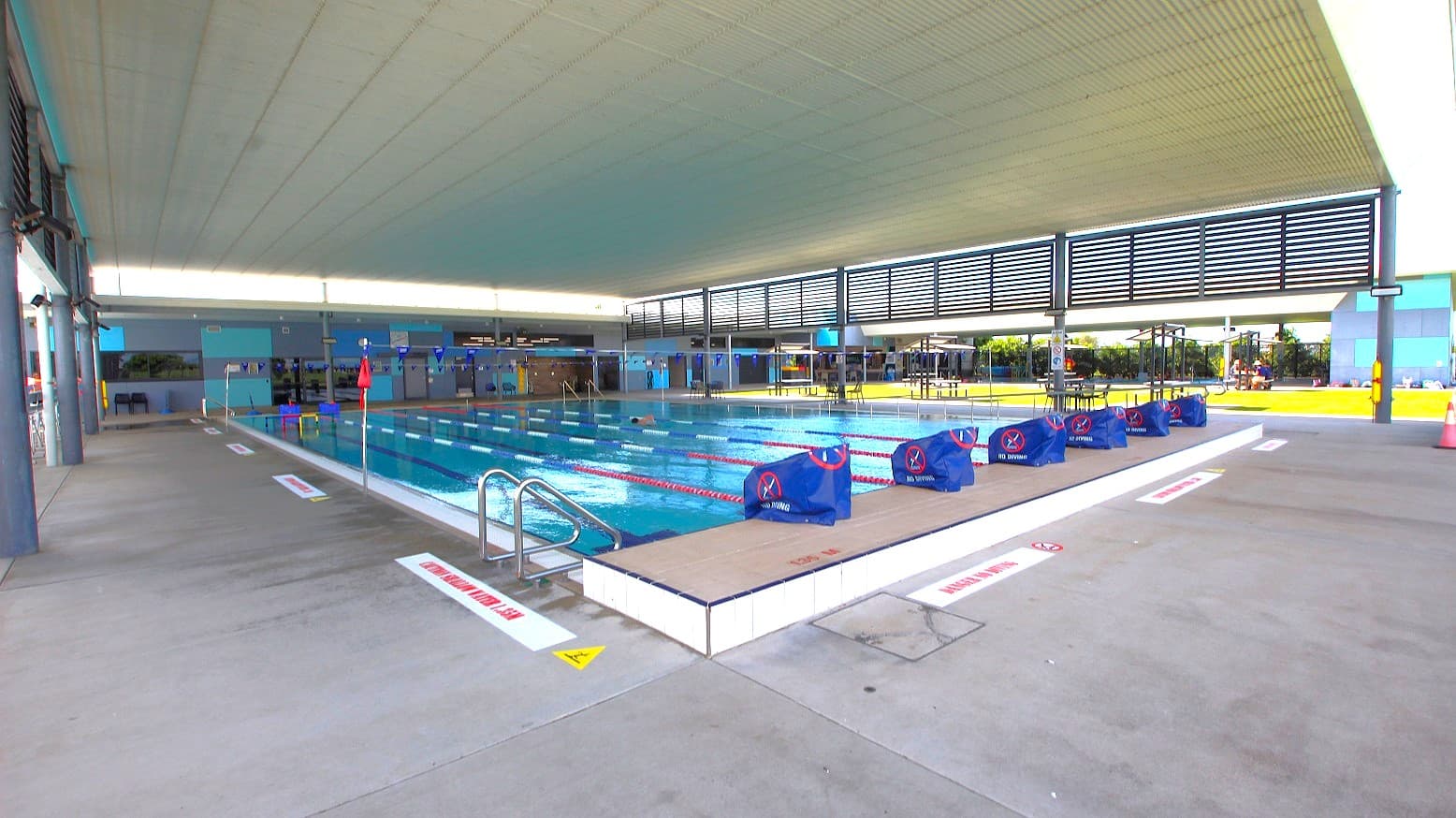 An indoor swimming pool with multiple lanes, separated by red and white lane dividers. Blue starting blocks are positioned at one end. The pool is surrounded by a concrete deck with safety signs. The ceiling is high and white, with slatted panels allowing natural light. On the right, there are tables and chairs on a grassy area visible through large windows. The left side has a walkway with structural columns.