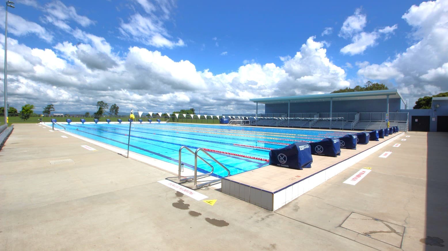 An outdoor swimming pool with multiple lanes, bordered by a concrete deck. On the right, there are covered seating areas with blue covers on the lane dividers. The left side shows an open area with a few trees in the distance. The pool has metal ladders for entry and exit. The sky is filled with scattered clouds.