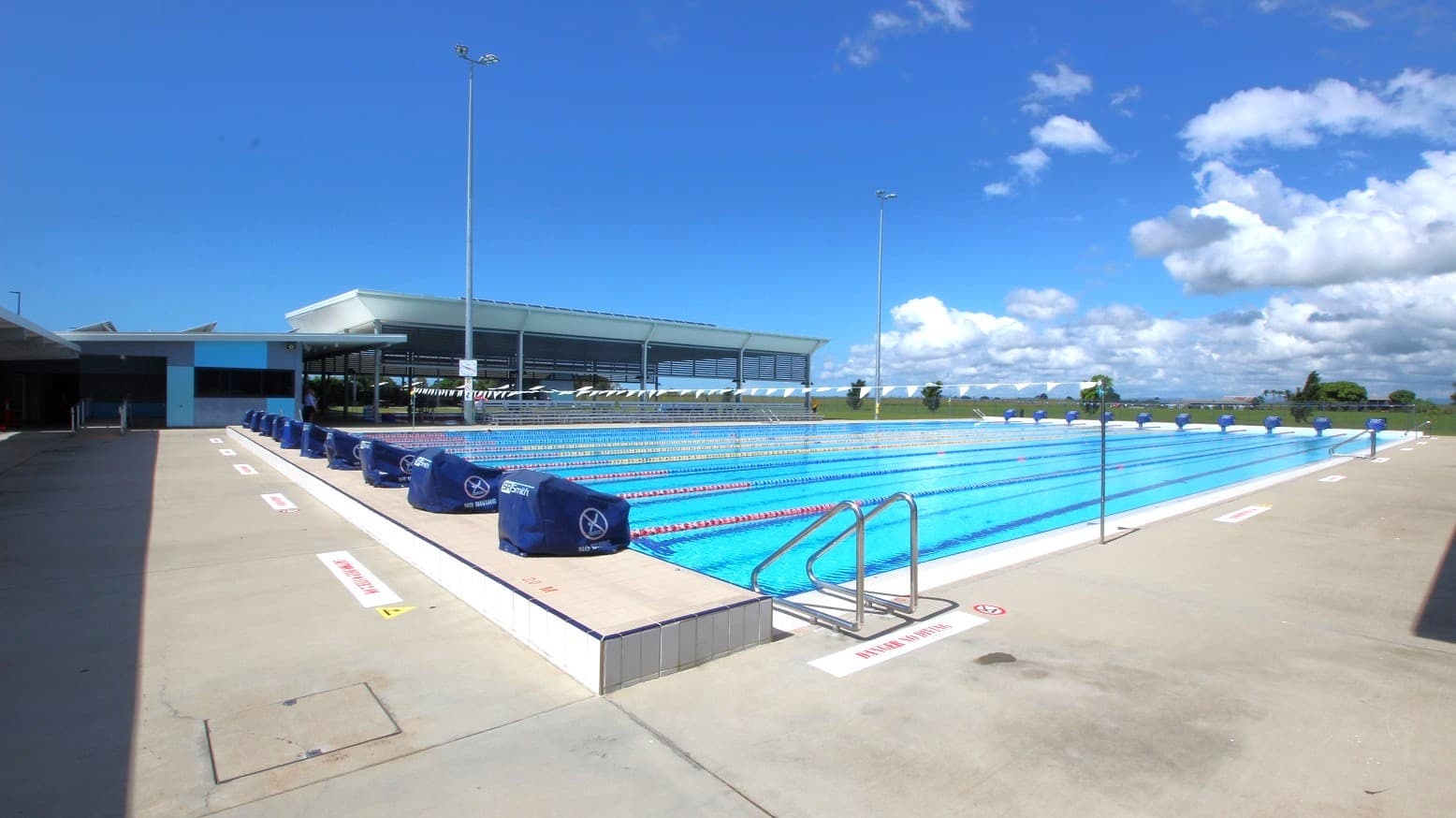 An outdoor swimming pool with multiple lanes, each marked by lane dividers. On the left, there is a shaded walkway leading to a building with a flat roof. The pool deck is concrete, with metal handrails at the pool's edge. In the background, a low building with large windows and a flat roof is visible. The pool is surrounded by open space with some trees and a clear sky above.