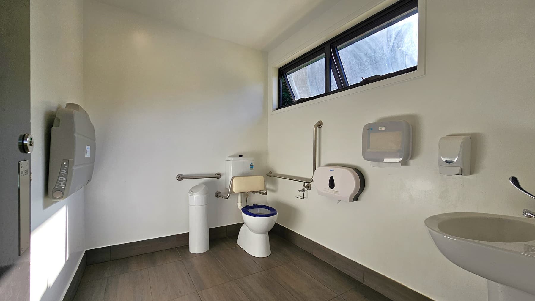 On the left, there is a white sink with a soap dispenser and a paper towel dispenser mounted on the wall above it. In the middle, there is a white toilet with a blue seat cover, a grab bar on the wall behind it, and a toilet paper dispenser to the side. On the right, there is a white waste bin and another grab bar on the wall. The floor is covered with grey tiles, and the ceiling is smooth and white with a recessed light.