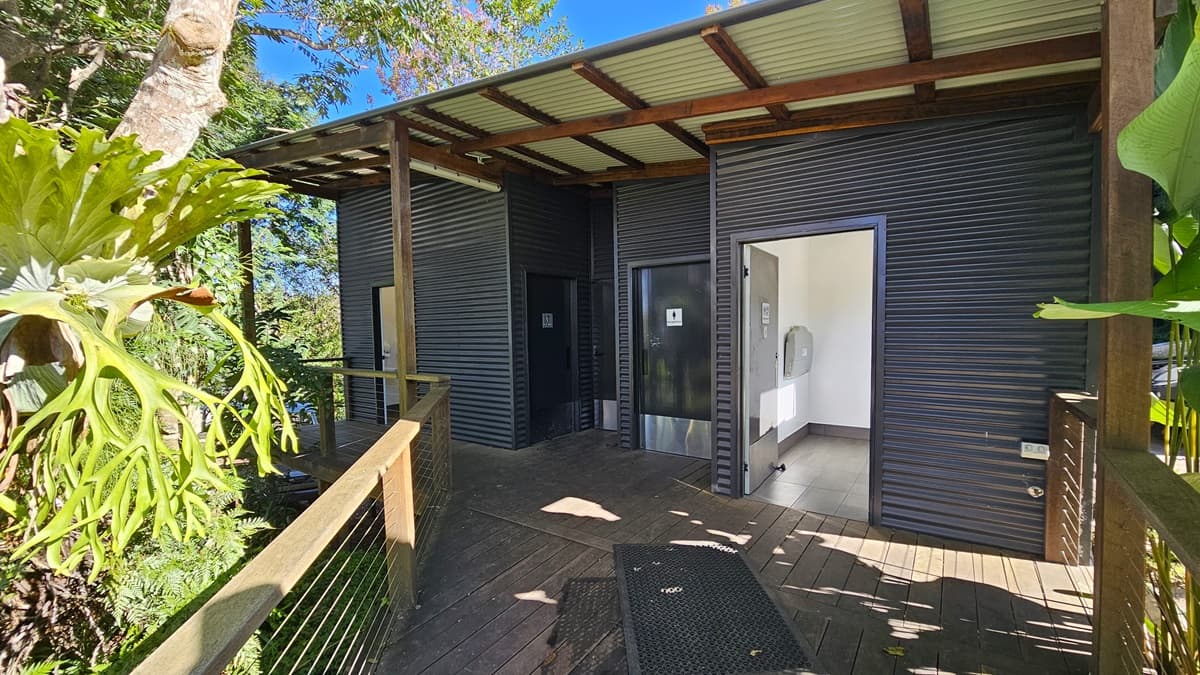 On the left, there is a large plant with broad leaves next to a wooden railing. In the middle, there are two dark grey metal structures with corrugated walls and open doorways, leading to interior spaces with light-coloured walls and tiled floors. On the right, there is another section of the dark grey metal structure with a small window. The floor is made of wooden planks, and the ceiling is a corrugated metal roof supported by wooden beams.