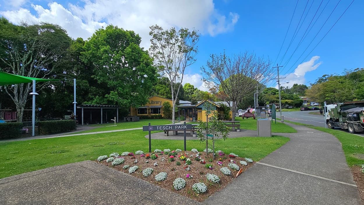 A grassy area with a wooden picnic table and benches on a concrete path in the foreground. A leafless tree stands nearby. In the middle, a yellow building with a sign reading "Library" is partially obscured by trees. To the right, a road with parked cars and a person walking. Power lines run alongside the road. Lush green trees are in the background.