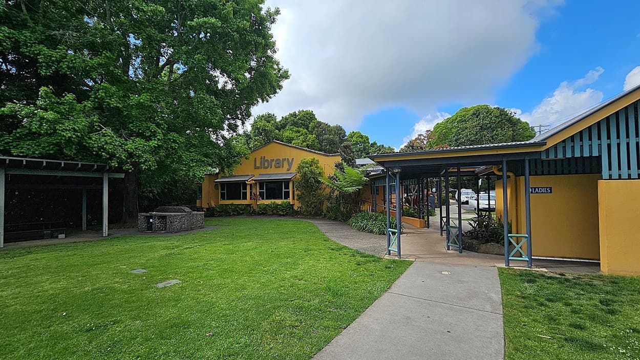 A yellow building with a sign reading "Library" is in the centre, surrounded by lush green trees and grass. To the right, there's a covered walkway leading to a building marked "Ladies". A paved path runs through the grass, and a few parked cars are visible in the background. On the left, there's a shaded area with a stone structure, possibly a barbecue or seating area.