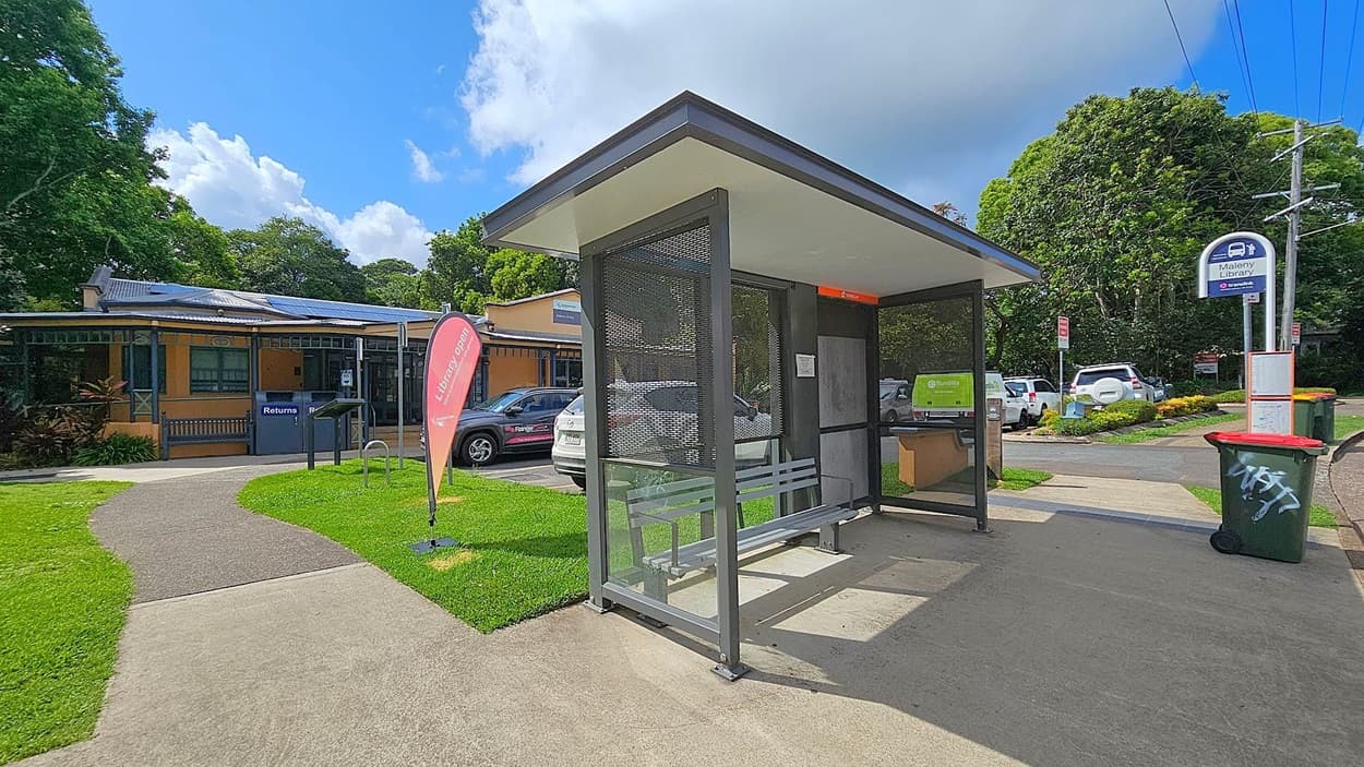 A bus stop with a metal bench and a shelter is on the right. A sign reads "Maleny Library." A red bin is nearby. On the left, a building with solar panels and a "Library Open" flag is visible. Several parked cars are in the background, surrounded by lush green trees. A pathway leads from the bus stop to the building.