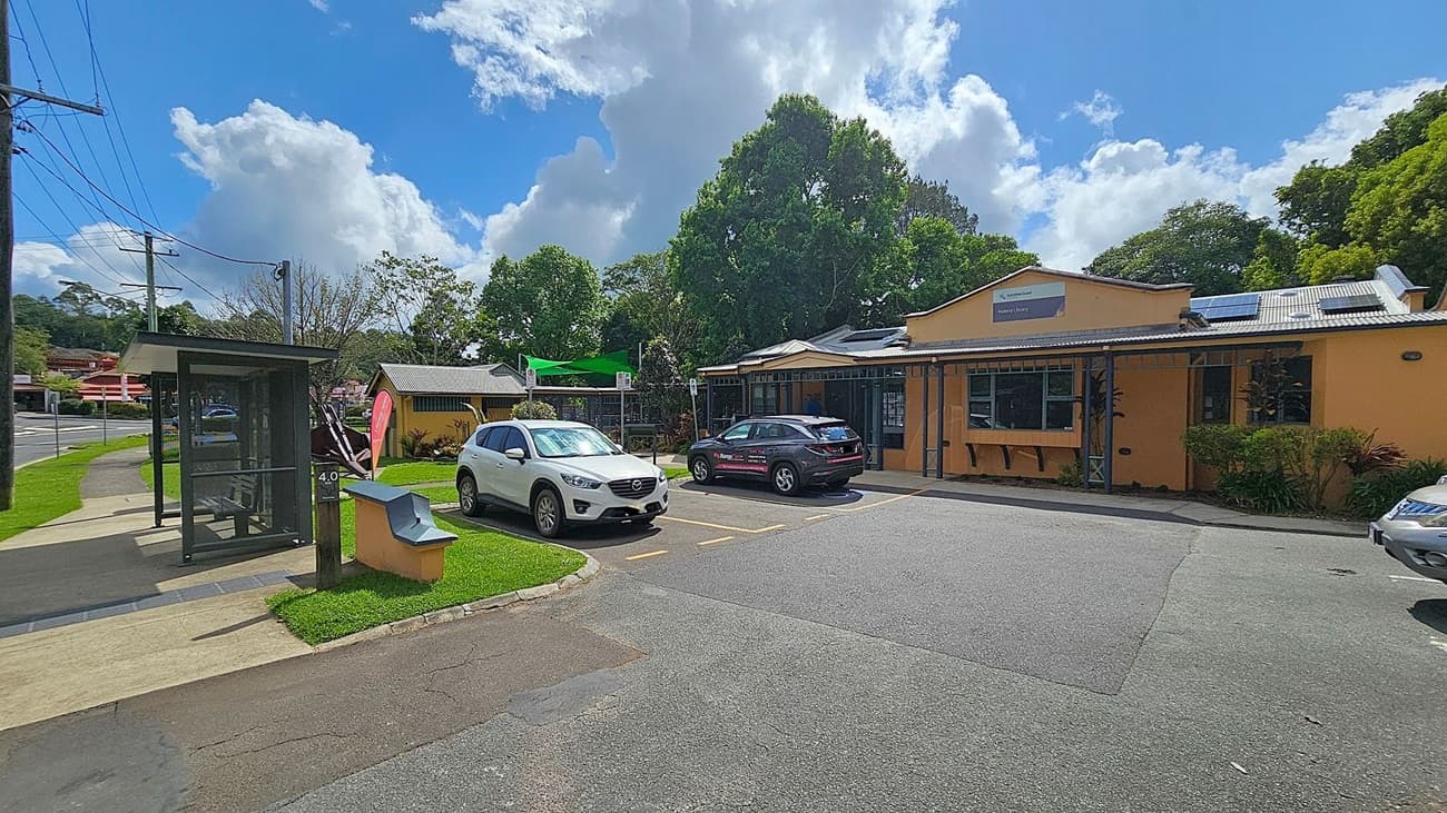 A street scene with a bus shelter on the left, next to a footpath and grassy area. Two cars are parked in front of a yellow building on the right, which has a sign indicating it's a library. The building is surrounded by trees and bushes. The sky is partly cloudy.