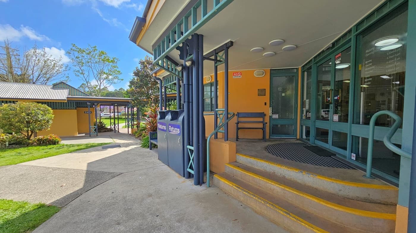 A building entrance with a yellow and green colour scheme. On the right, there are glass doors with operating hours listed and a "No Smoking" sign above. Two blue bins marked "Returns" are near the steps. The steps have yellow edges and a metal handrail. On the left, a path leads to another building with a corrugated metal roof. The area is landscaped with bushes and trees, and the ground is a mix of grass and concrete.