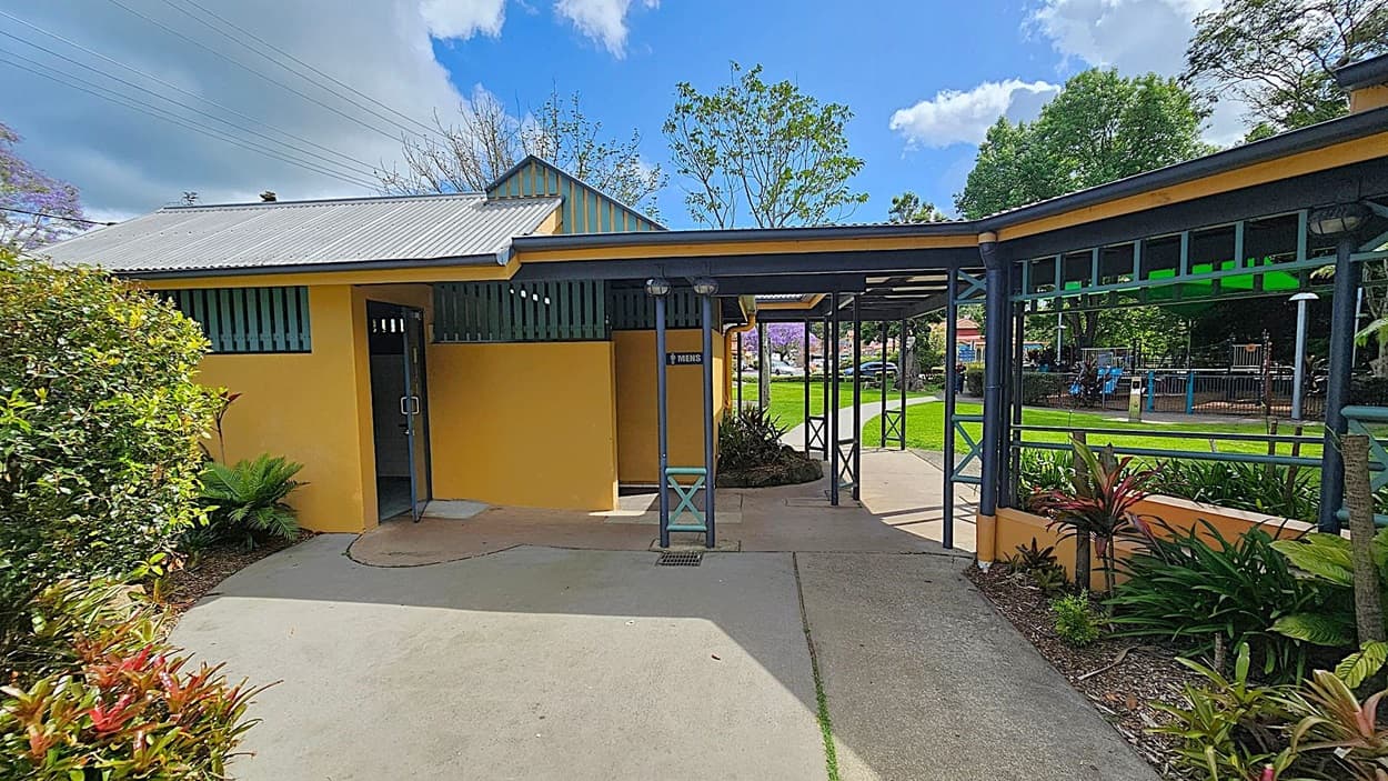 A yellow building with a corrugated metal roof is on the left, featuring a door marked "Men's". A covered walkway with green metal supports extends to the right, leading to a grassy area with trees and a playground in the background. The foreground has a concrete path bordered by various plants and shrubs.