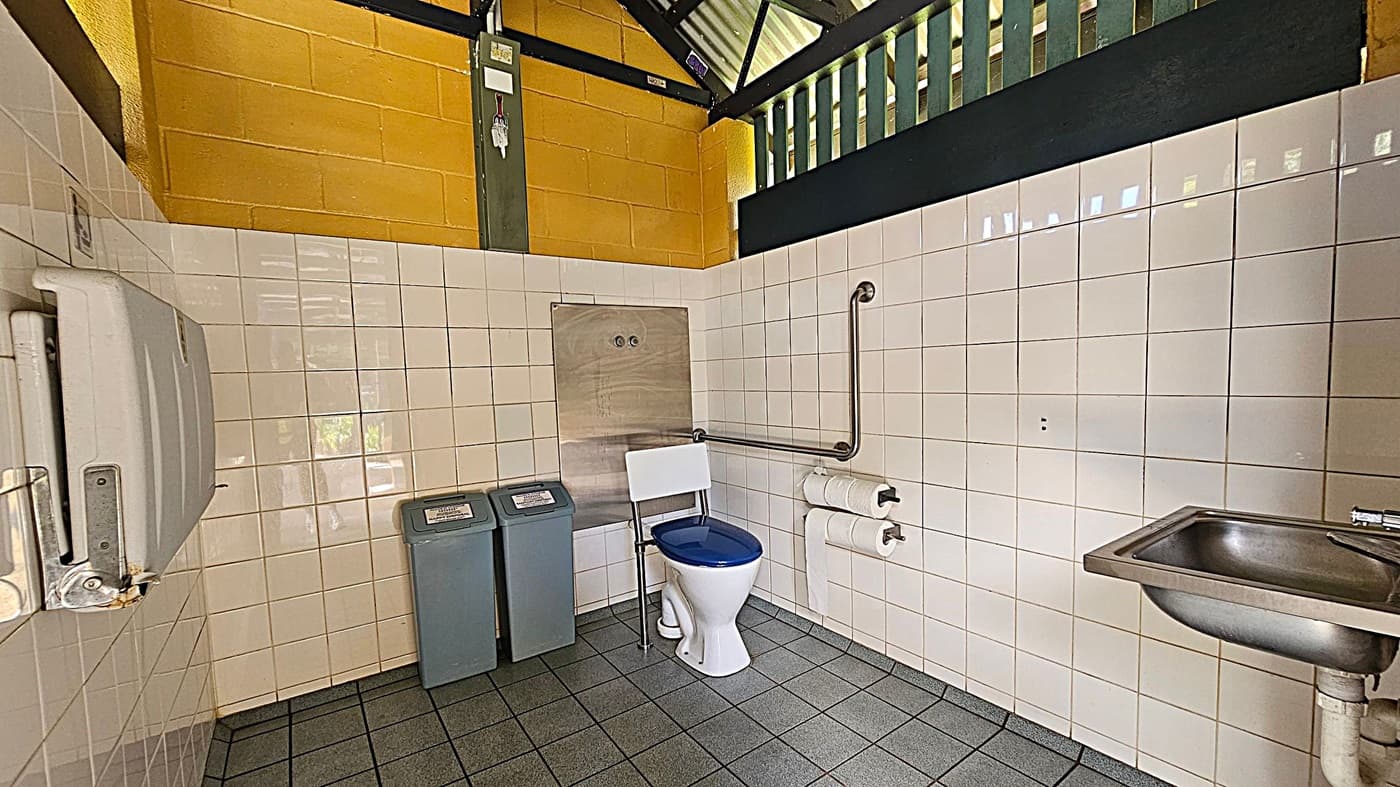 A public toilet with white tiled walls and a green-tiled floor. On the left, there's a hand dryer. In the centre, a toilet with a blue seat is positioned next to a stainless steel panel and two waste bins. A grab rail is mounted on the wall beside the toilet, with two toilet roll holders beneath it. On the right, a small stainless steel sink is attached to the wall. The upper wall is painted yellow, and the ceiling has a slatted design allowing natural light.