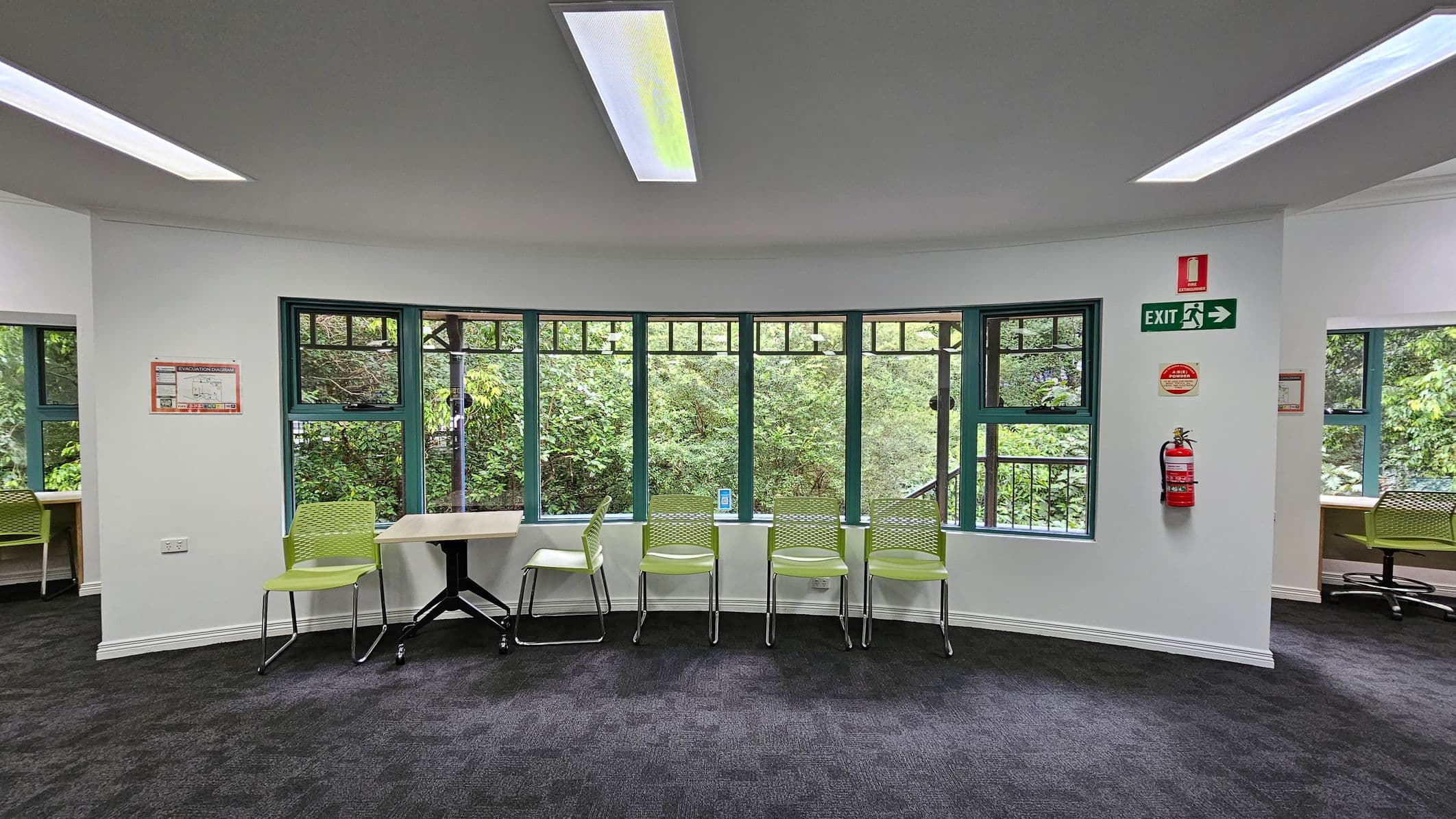 A library interior with dark carpeted flooring and a white ceiling featuring recessed lighting. In the foreground, there are several bookshelves filled with books on wheels. On the left, a desk with computers and colourful chairs is positioned near a green door. In the background, large windows and glass doors allow natural light to enter. A small TV is mounted on the wall to the right, above additional bookshelves.