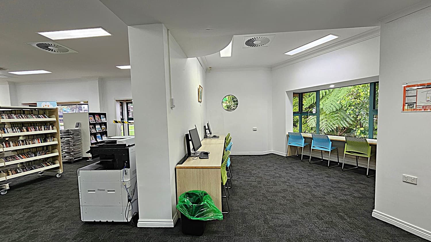 A library interior with a dark carpeted floor. On the left, there are bookshelves filled with books and a printer. In the middle, a row of computers on a wooden desk with colourful chairs. A green bin is beneath the desk. On the right, a large window with a view of lush greenery outside, and more colourful chairs lined up against a counter. The ceiling has recessed lighting and air vents.