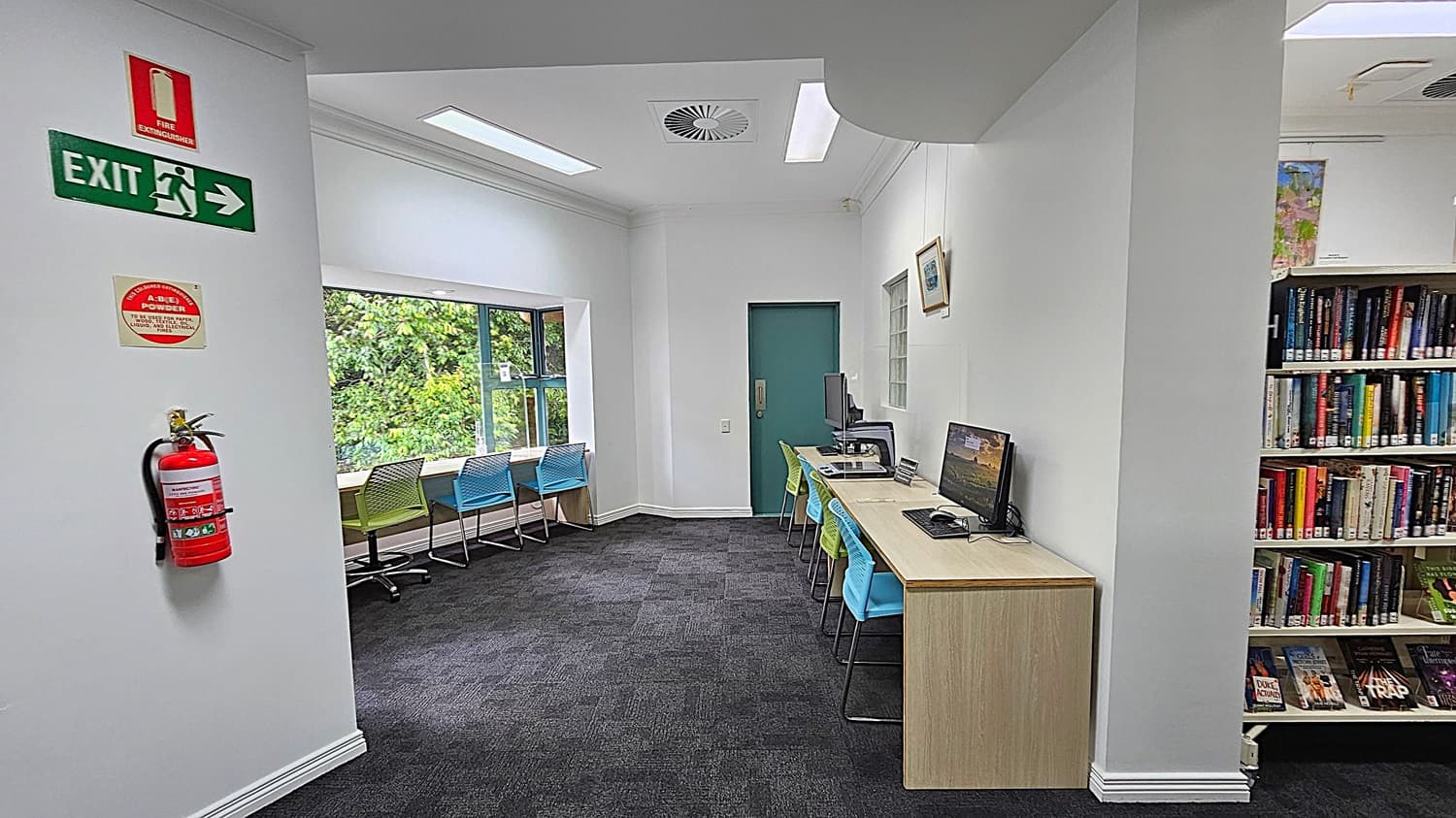 A library interior with a dark carpeted floor. On the left, a fire extinguisher and exit sign are mounted on the wall. In the middle, a row of colourful chairs faces a large window with a view of greenery. On the right, a long desk with computers and chairs is positioned against the wall. Bookshelves filled with books are visible further to the right. The ceiling has recessed lighting and a ventilation duct.