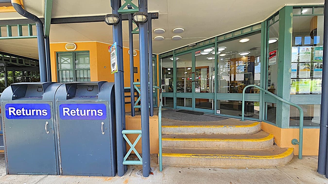 On the left, there are two blue book return bins labelled "Returns" with handles. In the middle, a set of concrete steps with yellow edges leads up to a glass entrance with green frames. The entrance has a rubber mat and several signs on the glass. The building has an orange wall and a white ceiling with circular lights. A green metal railing runs alongside the steps.