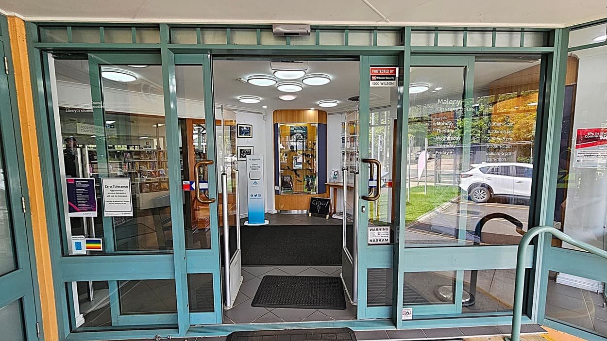Glass entrance doors with green frames lead into a building. On the left door, there are several notices, including a "Zero Tolerance" sign and a rainbow sticker. The right door displays opening hours. Inside, a display case and a small table are visible. The floor is tiled with black mats, and circular lights are on the ceiling. Outside, a white car is parked on the right, and greenery is visible through the glass.