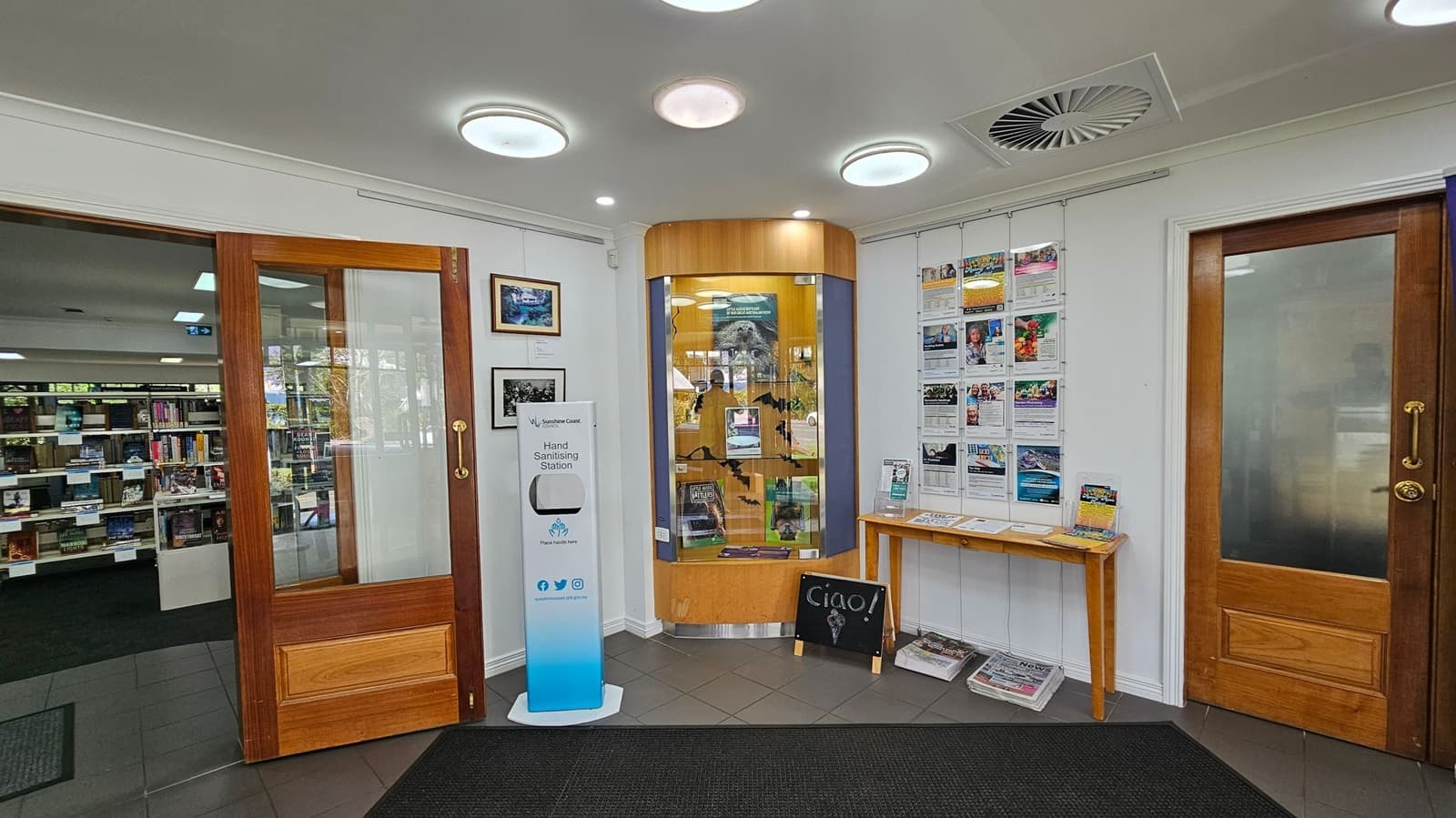 A library entrance area with wooden doors on either side. The left door is open, revealing bookshelves inside. In the centre, a glass display case contains various items. A hand sanitising station stands to the left. On the right, a wall-mounted rack holds brochures above a small table with more pamphlets and newspapers. The floor is tiled, and the ceiling has circular lights and a vent.