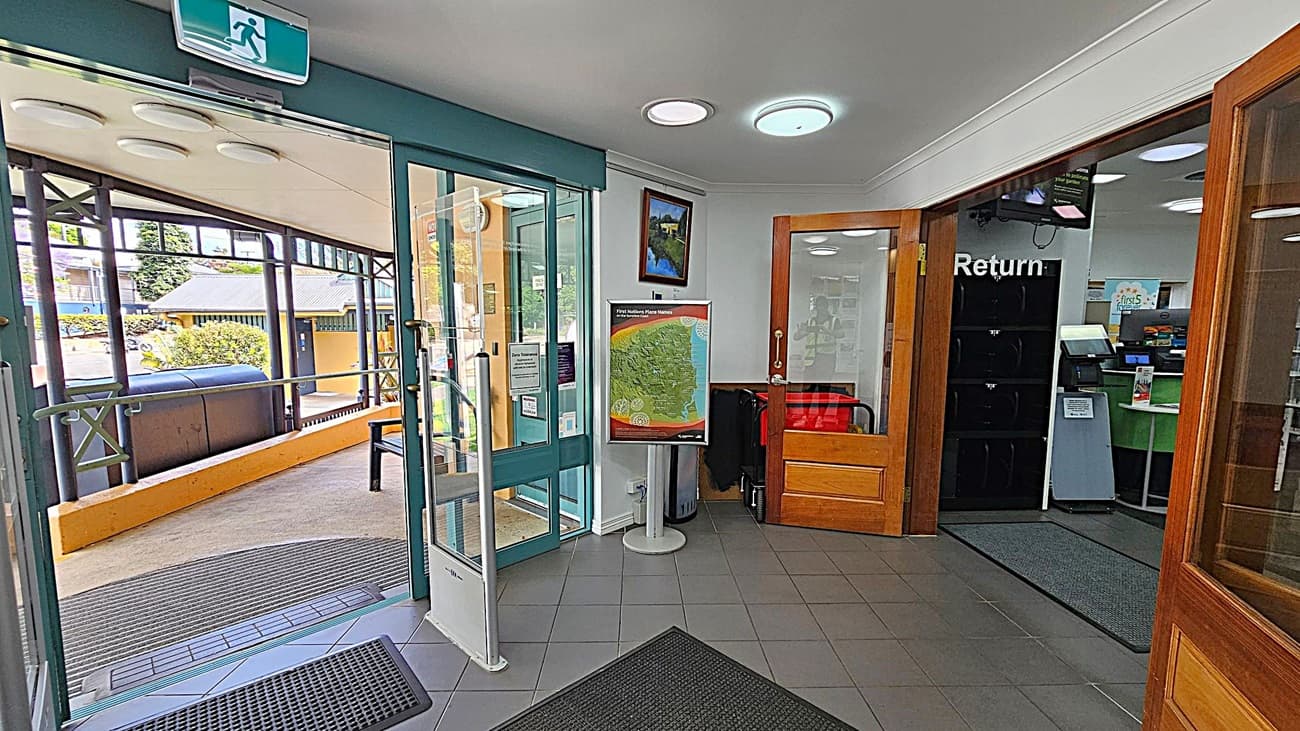 A library entrance area with a glass door on the left leading outside, where a covered walkway and some buildings are visible. Inside, a map on a stand and a framed picture are on the wall. To the right, there's a wooden door with a "Return" sign above it, leading to a room with a counter and computer screens. The floor is tiled with grey mats, and the ceiling has circular lights.