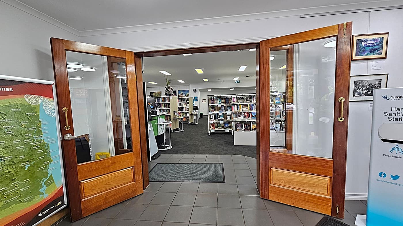Wooden double doors with glass panels open into a library. On the left, a large map is displayed. On the right, a hand sanitising station and framed pictures are visible. Inside, bookshelves filled with books are arranged neatly, and a counter with a computer is on the left. The floor is tiled near the entrance and carpeted inside. The ceiling has recessed lighting.