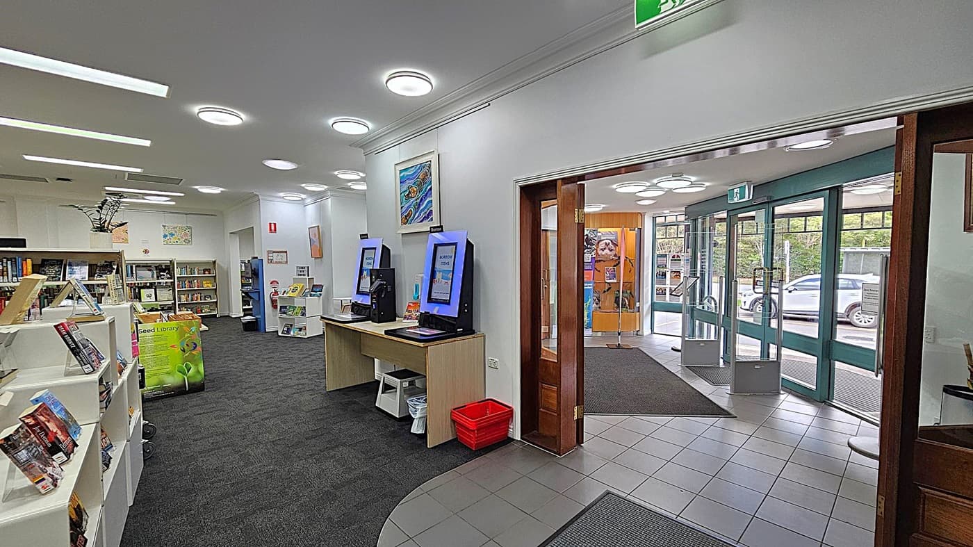 A library interior with bookshelves on the left filled with books. In the centre, there are two self-service kiosks on a wooden desk. The floor is carpeted in the library area and tiled near the entrance. On the right, glass doors lead outside, with a view of parked cars. The ceiling has circular lights, and there are various posters and artworks on the walls.