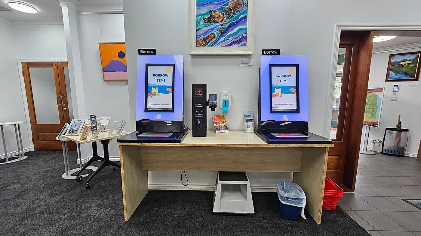 Two self-service kiosks with screens displaying "Borrow Items" are on a wooden desk. Between them is a payment terminal and a hand sanitiser dispenser. Above, a colourful painting is mounted on the wall. To the left, a small table holds books, and a wooden door is visible. On the right, another wooden door leads to a tiled area with a map and a waste bin. The floor is carpeted in dark grey.