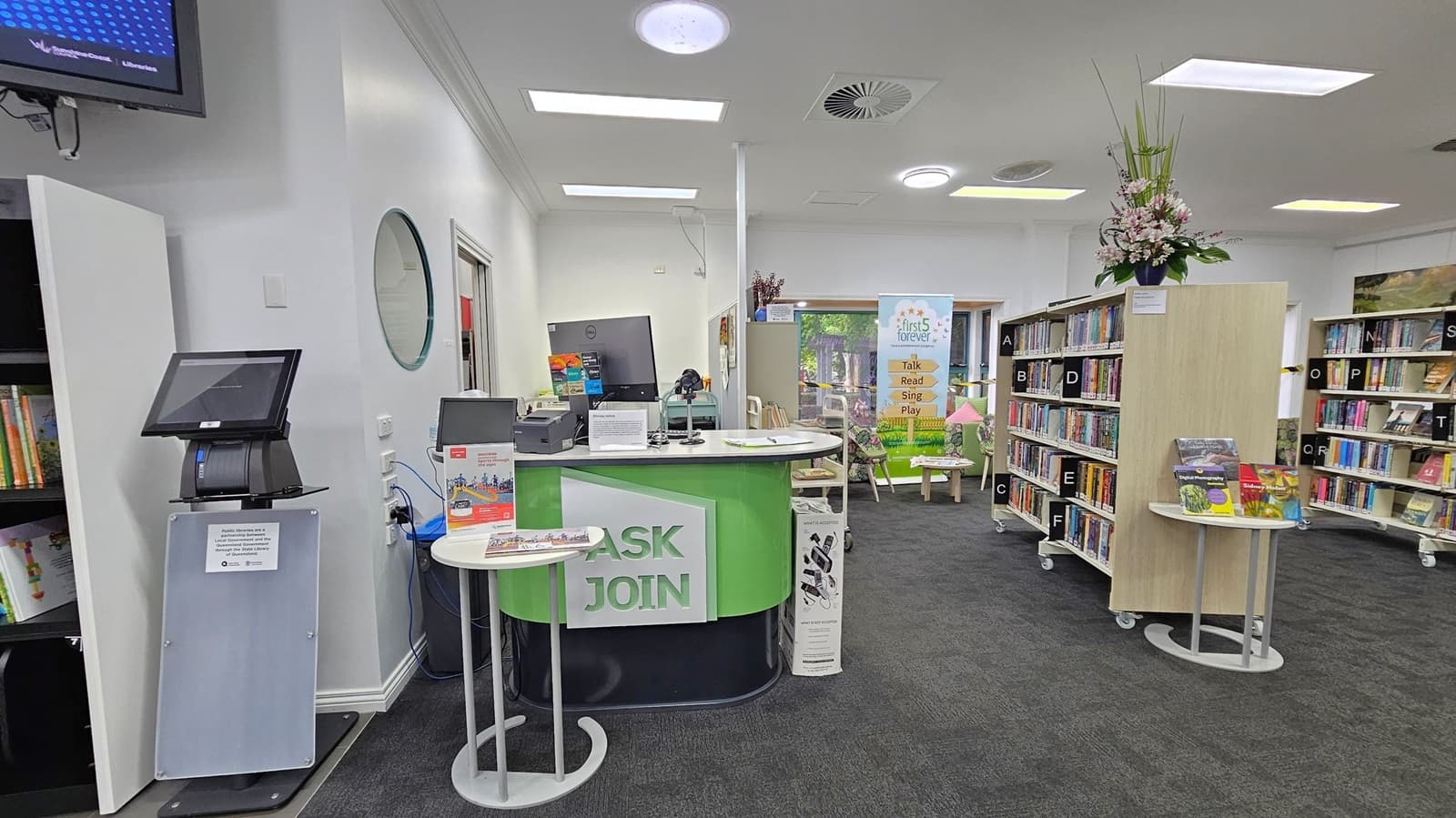 A library interior with a black carpeted floor. On the left, there's a self-service kiosk with a touchscreen. Next to it is a green and white desk with "ASK JOIN" signage, featuring a computer and brochures. In the middle, a banner reads "first 5 forever" with a small seating area behind it. On the right, bookshelves labelled with letters hold various books. A small round table displays more books, and a tall floral arrangement sits atop a shelf. The ceiling has recessed lighting and air vents.