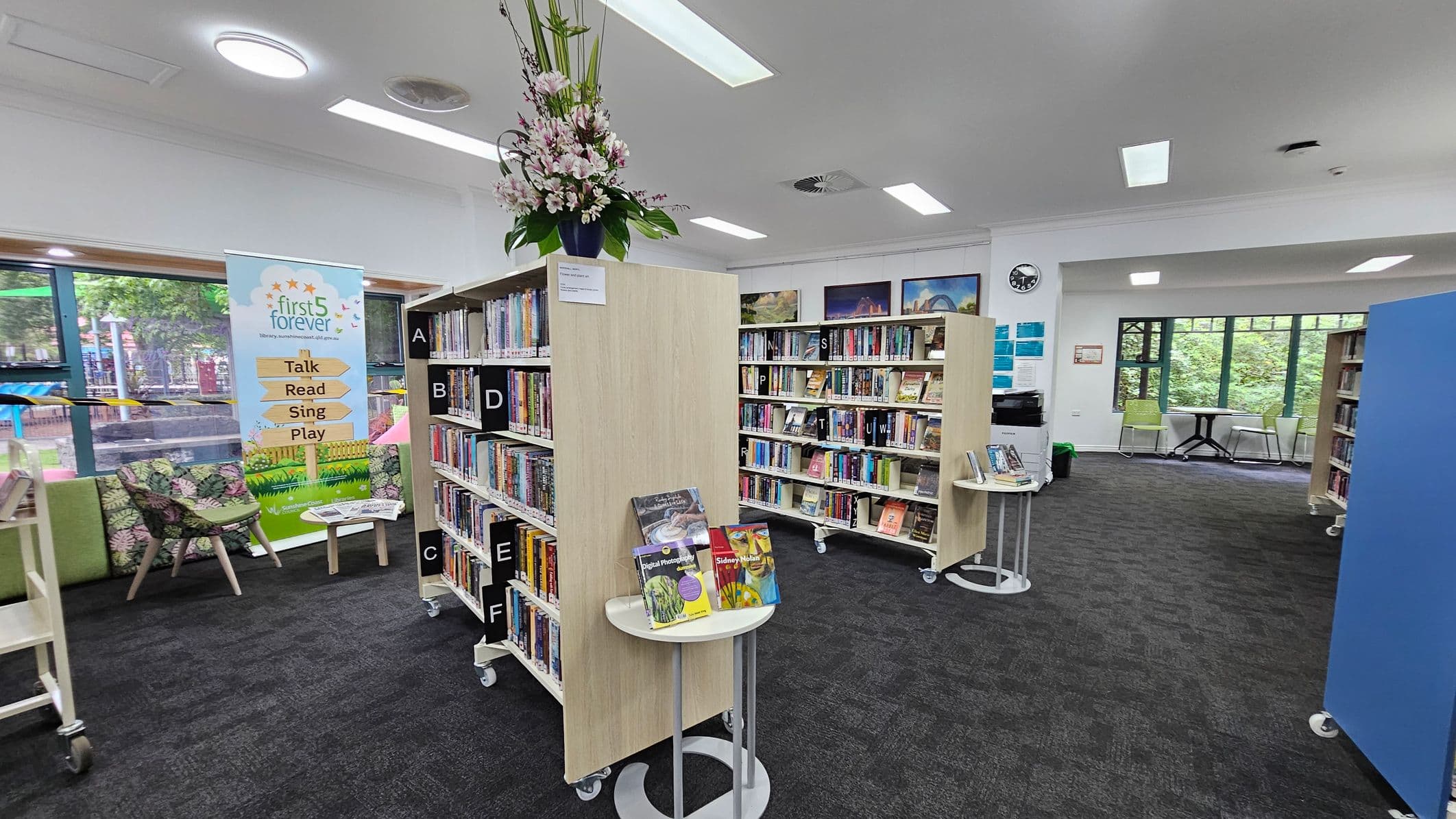 A library interior with several bookshelves on wheels filled with books. The left side has a large window with green frames, two green chairs, and a small round table. The middle features bookshelves with a nature-themed panel showing a waterfall and greenery. The right side has more bookshelves and a display stand with books. The floor is carpeted in dark grey, and the ceiling is white with rectangular lights.