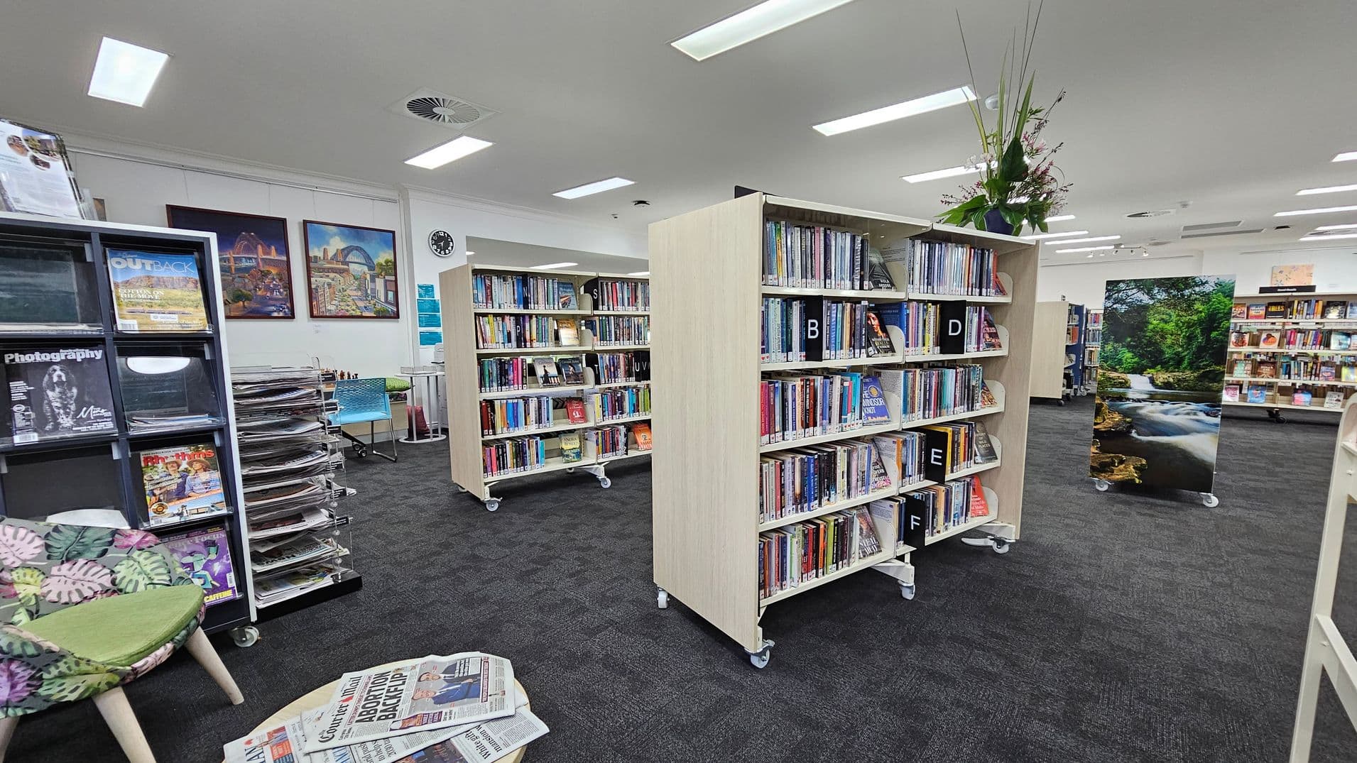 A library interior with bookshelves on wheels filled with books, labelled with letters. On the left, a magazine rack displays various titles, and a chair with a leaf-patterned cushion is nearby. Newspapers are spread on a small table. In the background, colourful paintings and a clock adorn the wall. A large photograph of a forest stream is positioned between the shelves. The floor is carpeted in dark grey, and the ceiling has recessed lighting.