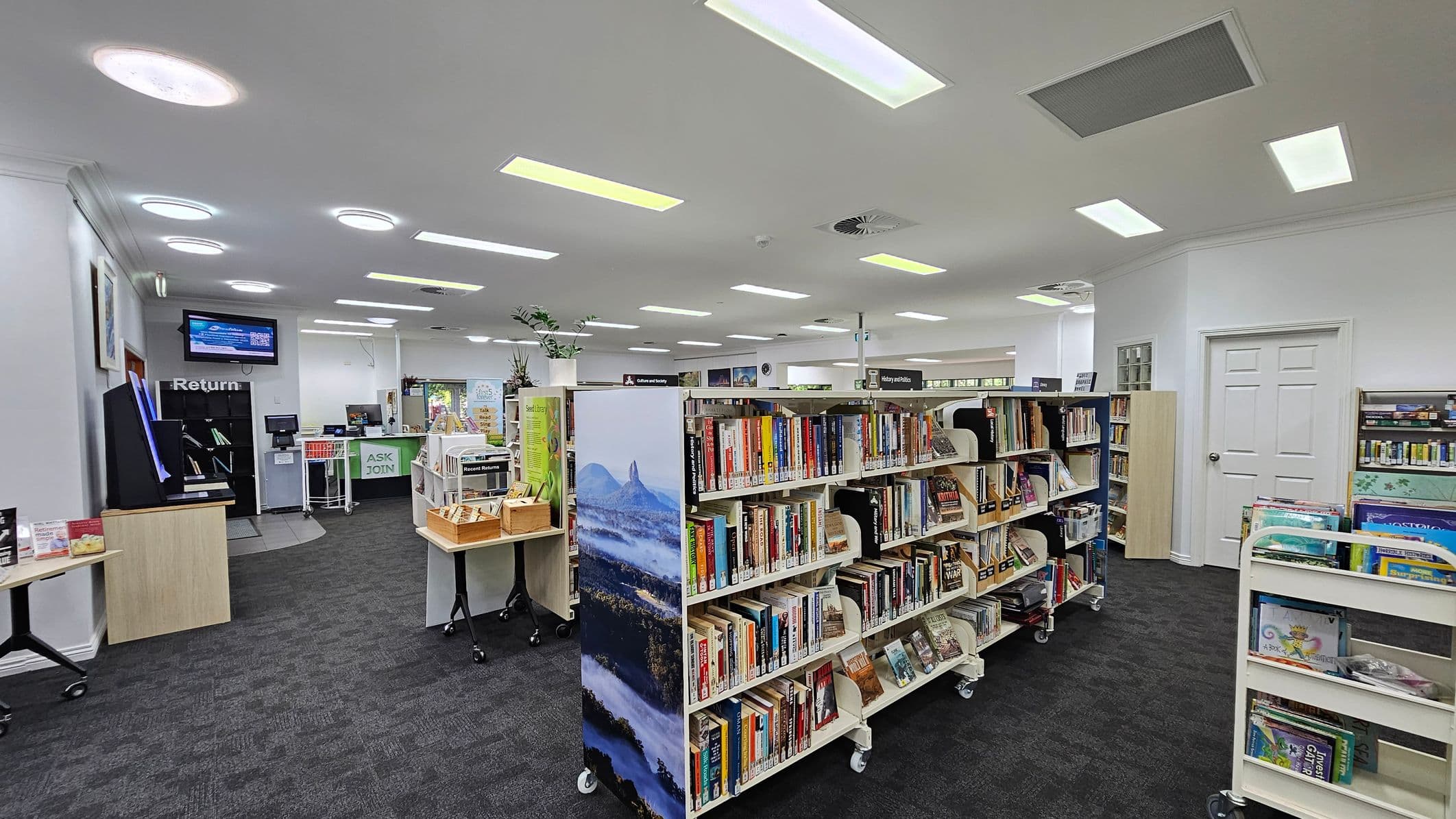 A library interior with bookshelves filled with books in the centre. On the left, there's a book return station and a digital screen above it. To the right, a white door and more bookshelves are visible. The floor is carpeted in dark grey, and the ceiling is white with recessed lighting and circular fixtures. In the background, a counter with a sign reading "ASK JOIN" is present, along with various displays and plants.
