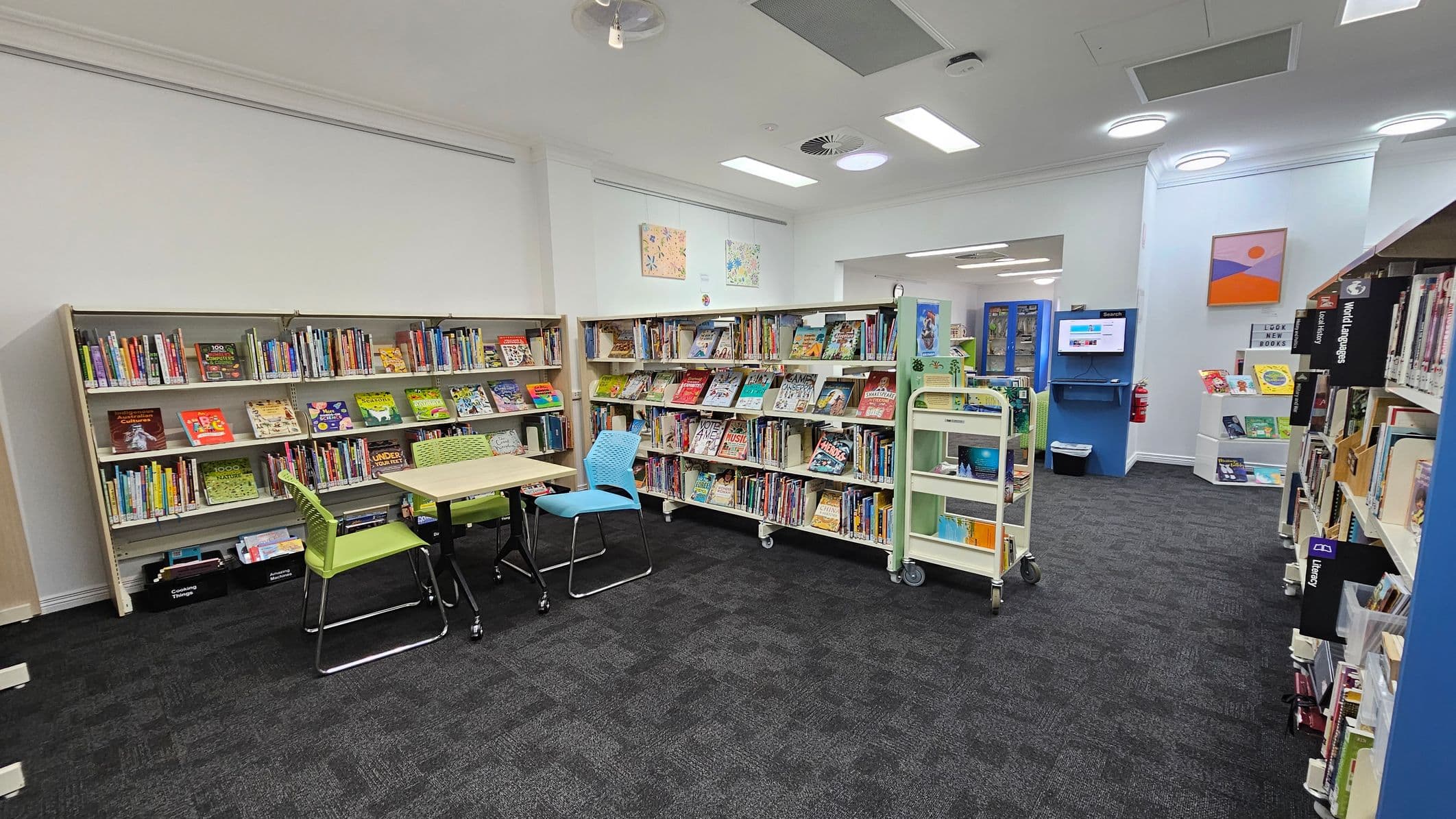 A library interior with bookshelves lining the left and right walls, filled with colourful children's books. In the centre, there is a small table with green and blue chairs. The floor is carpeted in dark grey. On the right, a digital search station is mounted on a blue wall, with a fire extinguisher nearby. The ceiling has recessed lighting and ventilation panels. Artwork is displayed above the shelves.