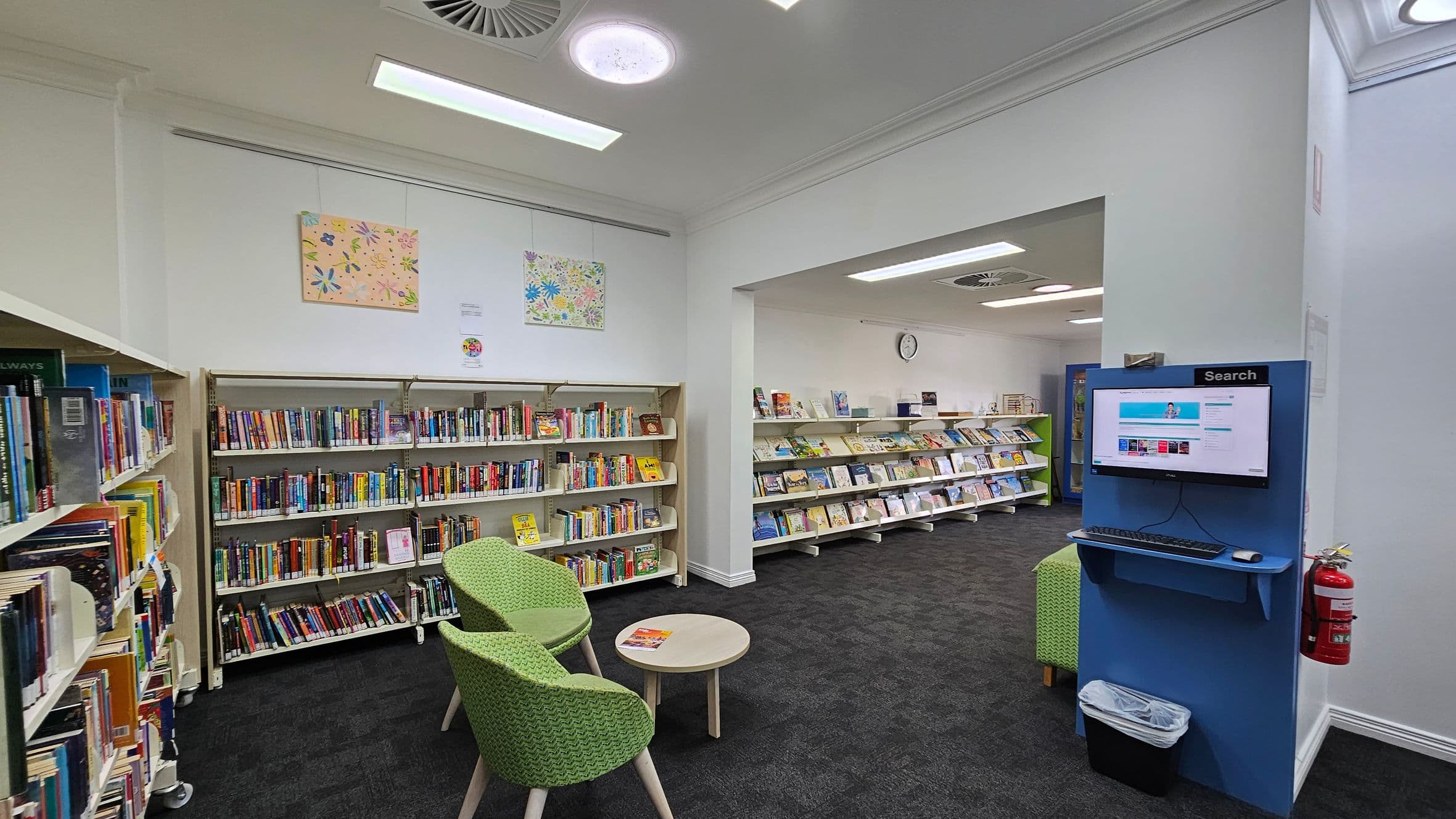A library interior with bookshelves lining the left wall, filled with various books. Two green chairs and a small round table are in the foreground. On the right, a blue search station with a computer screen and keyboard is mounted on the wall, with a fire extinguisher below. The back wall features more bookshelves and a clock. The ceiling has circular lights and vents, and the floor is carpeted in dark grey. Artwork is displayed above the bookshelves.