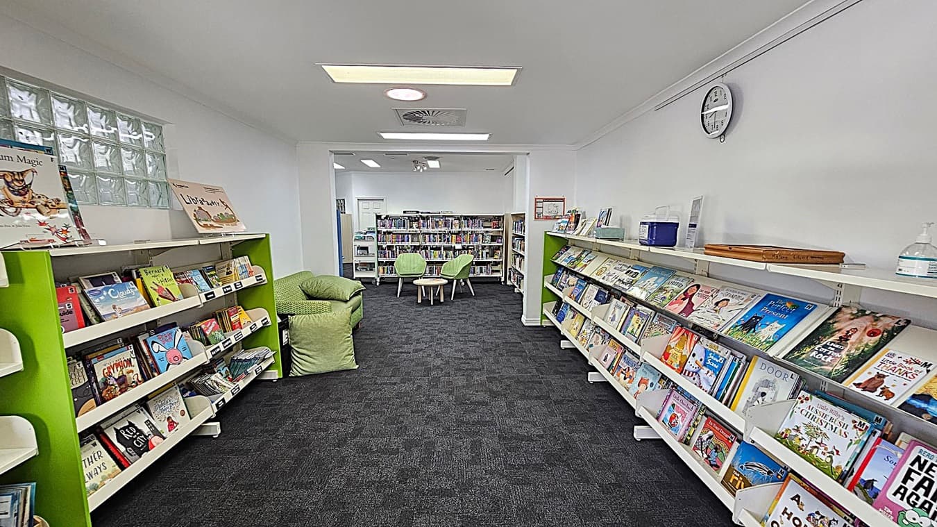 A library with a dark carpeted floor and white ceiling. On the left, there are green bookshelves filled with children's books. A green cushioned chair is nearby. On the right, more bookshelves display a variety of books, with a clock on the wall above. In the centre, two green chairs and a small table are positioned in front of more bookshelves. The room is well-lit with overhead lights.