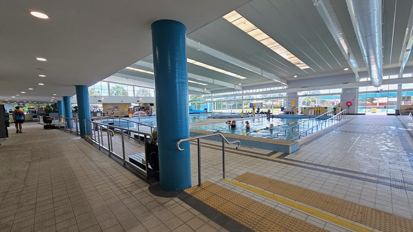 Access to the pool is via a ramp with handrails on the left, and on the right, there's a stepped entry with handrails and truncated domes. The Kids Leisure Pool and training Pool are on the far left, equipped with a ramp and handrail. To the right is the 25m lap pool with ladder access. A pool deck separates the two pools. The floor has matte grey tiles, and the ceiling features strips of transparent roofing, allowing natural sunlight and large aircon vents.