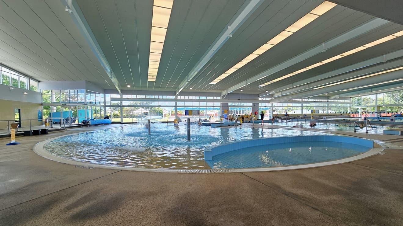 In the middle is the Leisure Pool with a lazy river ride, umbrella water features, and a walled toddler area. The pool is surrounded by an overflow grid. On the left, there is bench seating and a free-standing banner on the pool deck. To the right is the Program Pool equipped with handrails. The area is surrounded by glass, and the ceiling features strips of transparent roofing that allow natural sunlight.