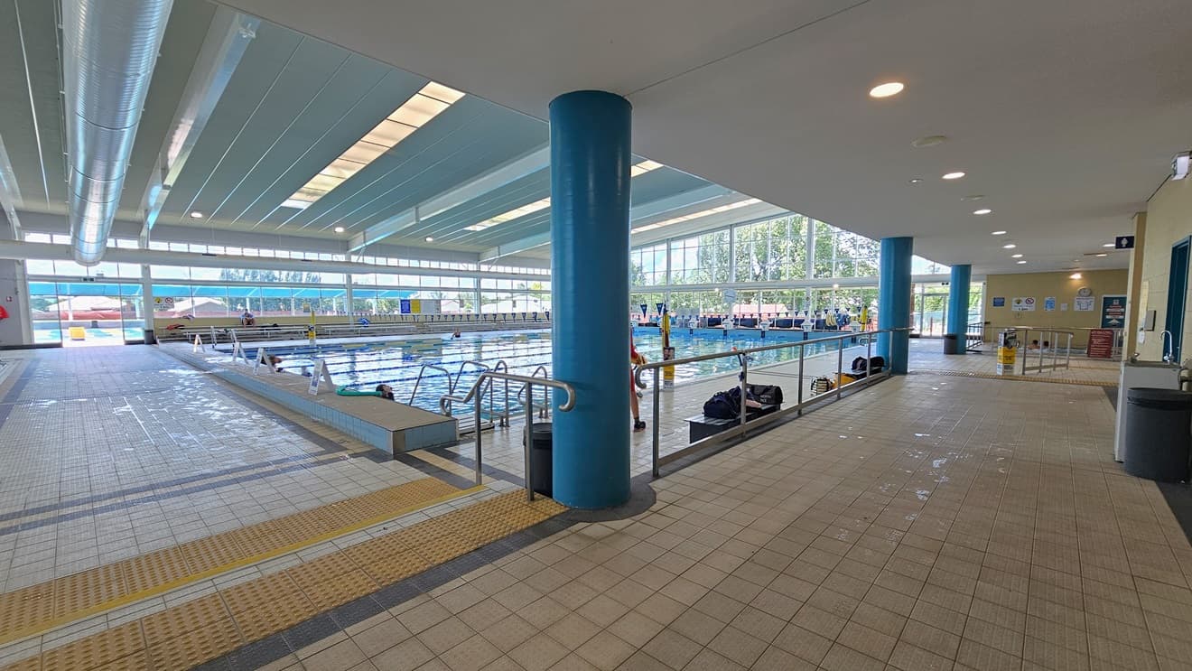 Pool access is via a left ramp with handrails and a right stepped entry with handrails and truncated domes. The 25m lap pool with ladder and handrail access is on the right. Freestanding banners adorn the pool deck. The floor features matte grey tiles, and the ceiling has strips of transparent roofing for natural sunlight, complemented by large aircon vents. The left corridor leads to the bathroom, where a drinking fountain, rubbish bins, and bench chairs are visible.