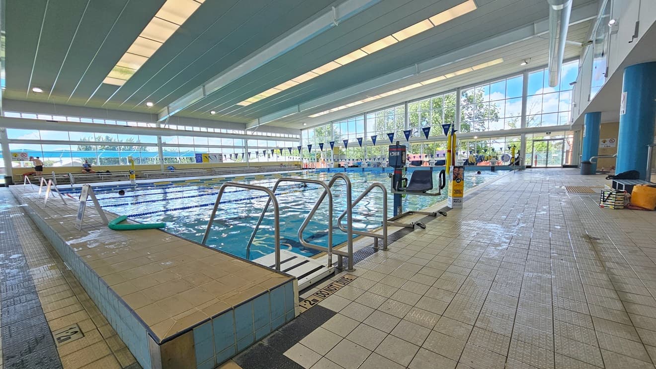 The 25m lap pool features a ladder and handrails, accompanied by an aquatic wheelchair and hoist in the corner. Free Standing banners and a green noodle hose decorate the left pool deck. Matte grey tiles cover the floor, and strips of transparent roofing in the ceiling provide natural sunlight, along with large aircon vents. Tiered seating is available on the far left, and bench chairs are positioned on the right.