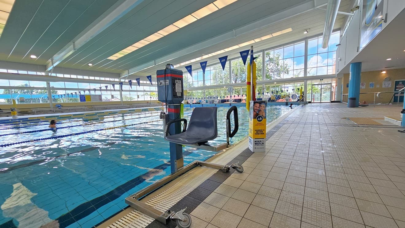 The 25m lap pool is equipped with a ladder and handrails, and the pool deck features an aquatic wheelchair and hoist. Floating ropes and blue bunting banners run across the pool. The high ceiling incorporates strips of transparent roofing, allowing natural sunlight, and large aircon vents. Tiered seating is available on the far left, and the floor is tiled in grey. A glass wall surrounds the area.