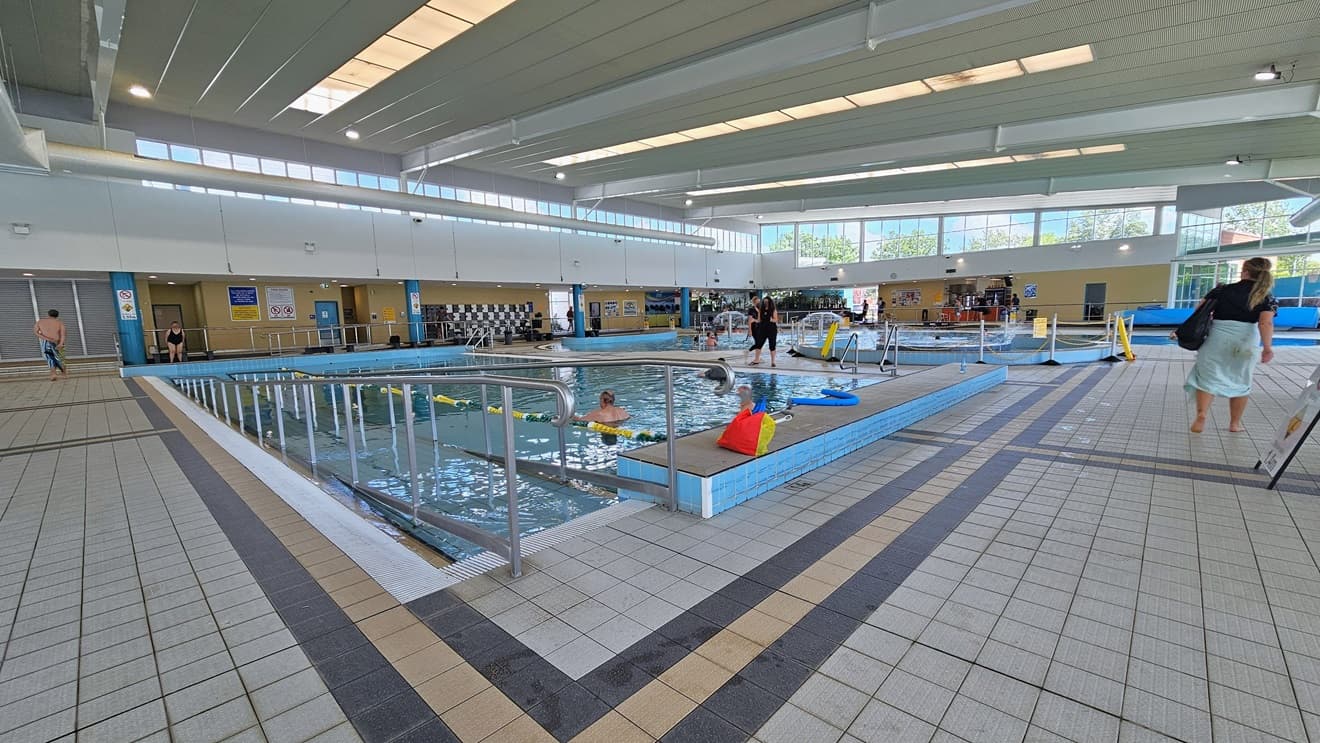 The Program Pool features a ramp with handrails for access, with floating ropes running across the pool. Belongings are visible on the raised pool deck. The Leisure Pool, located to the far right, is equipped with umbrella showers and water features, surrounded by rope bollards. Change places are situated on the opposite side of the pool. The floor has grey tiles, and guests are present in the pool. The surrounding upper wall is made of glass, providing a view of the outside.