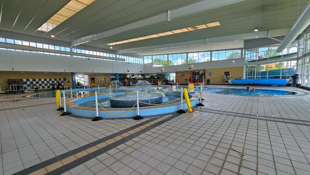 The Leisure Pool includes a lazy river ride with umbrella water features and a walled toddler area. A section of the pool has a rope bollard barricade. To the far left is the locker area. The floor features grey tiles, and guests are present in the pool. The surrounding upper wall is made of glass, providing a view of the outside. The high ceiling incorporates strips of transparent roofing, allowing natural sunlight, and large aircon vents.