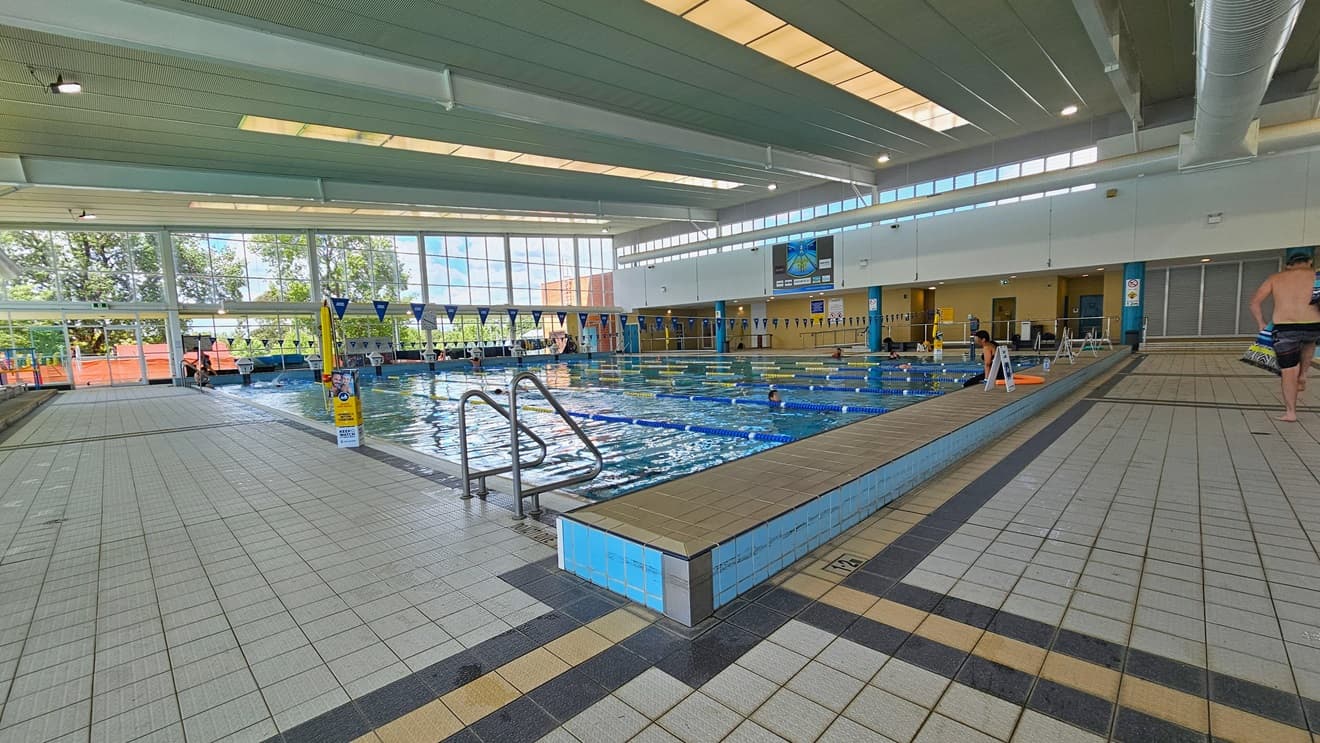 The pool is equipped with handrails, and banners are visible on the pool deck. Floating ropes and blue bunting run across the pool. The raised pool deck features free-standing banners. The left and right pool decks have grey tiles. The surrounding upper wall is made of glass, providing a view of the outside. The high ceiling incorporates strips of transparent roofing, allowing natural sunlight, and large aircon vents.