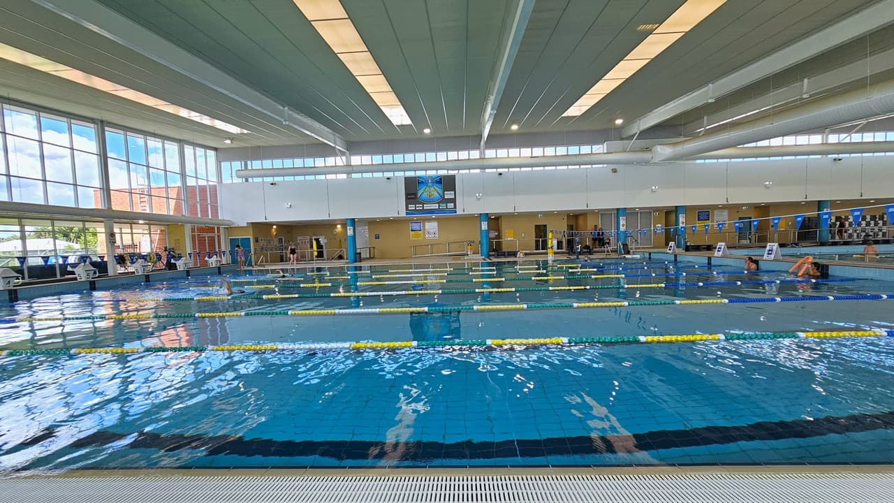 The pool is equipped with colorful floating ropes, and blue buntings run across it. The pool edge features an overflow grid. The raised pool deck has free-standing banners and diving boards. The surrounding upper wall is made of glass, providing a view of the outside with a large poster. The high ceiling incorporates strips of transparent roofing, allowing natural sunlight, and large aircon vents.