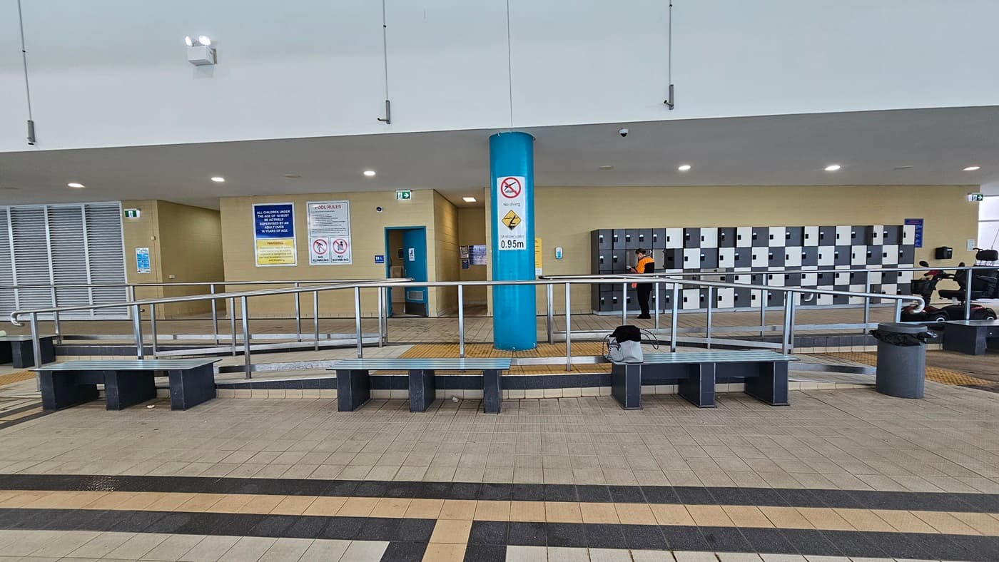 Bench chairs are placed near the ramp entrance, accompanied by rubbish bins. Behind, the locker area in alternating shades of black and white is located on the right. To the left are the accessible bathrooms, with safety posters and stickers affixed to the beige wall. The floor is adorned with cream tiles featuring a black and beige accent.